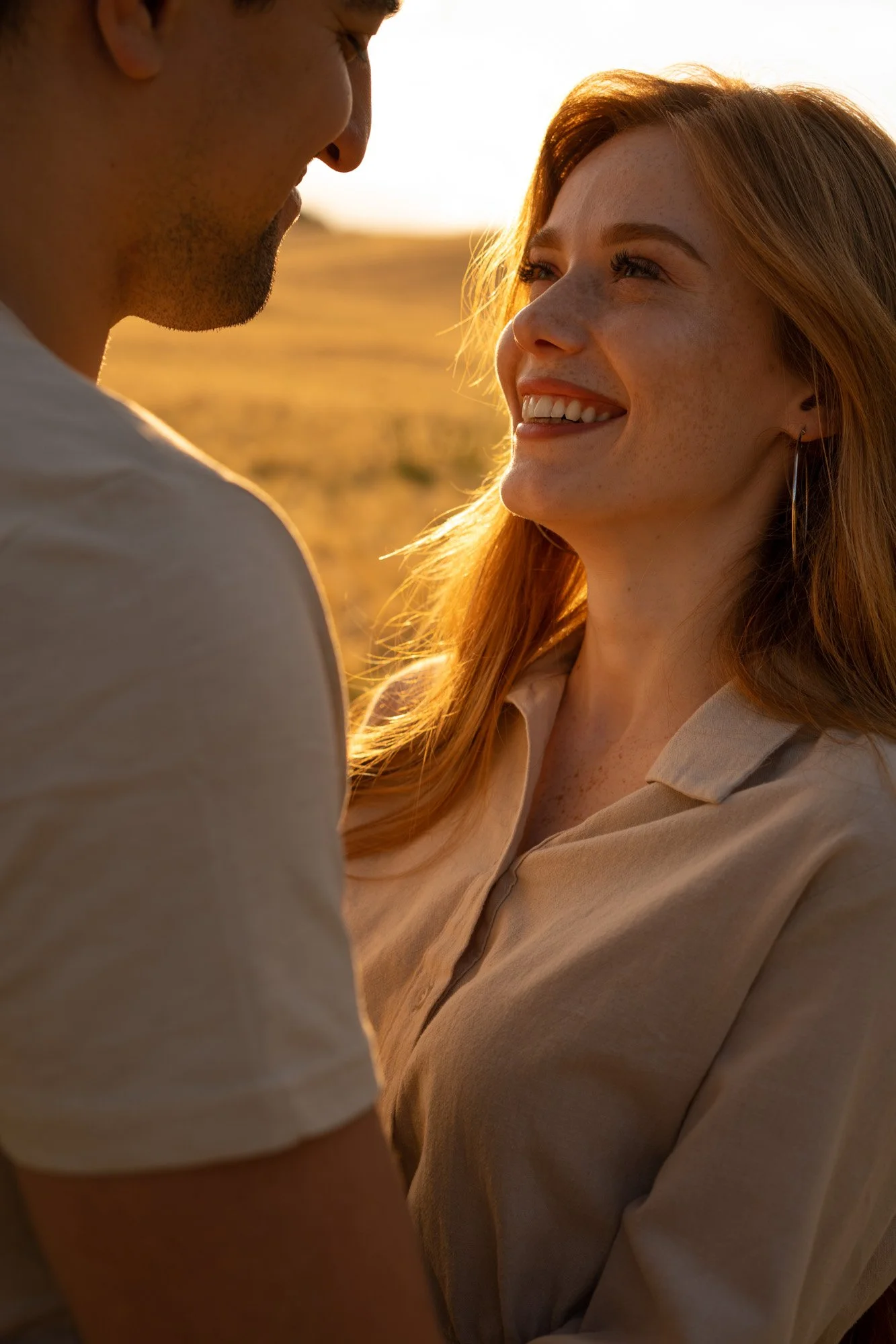 A woman with red hair smiling and looking into the eyes of a man, both outdoors during golden hour.