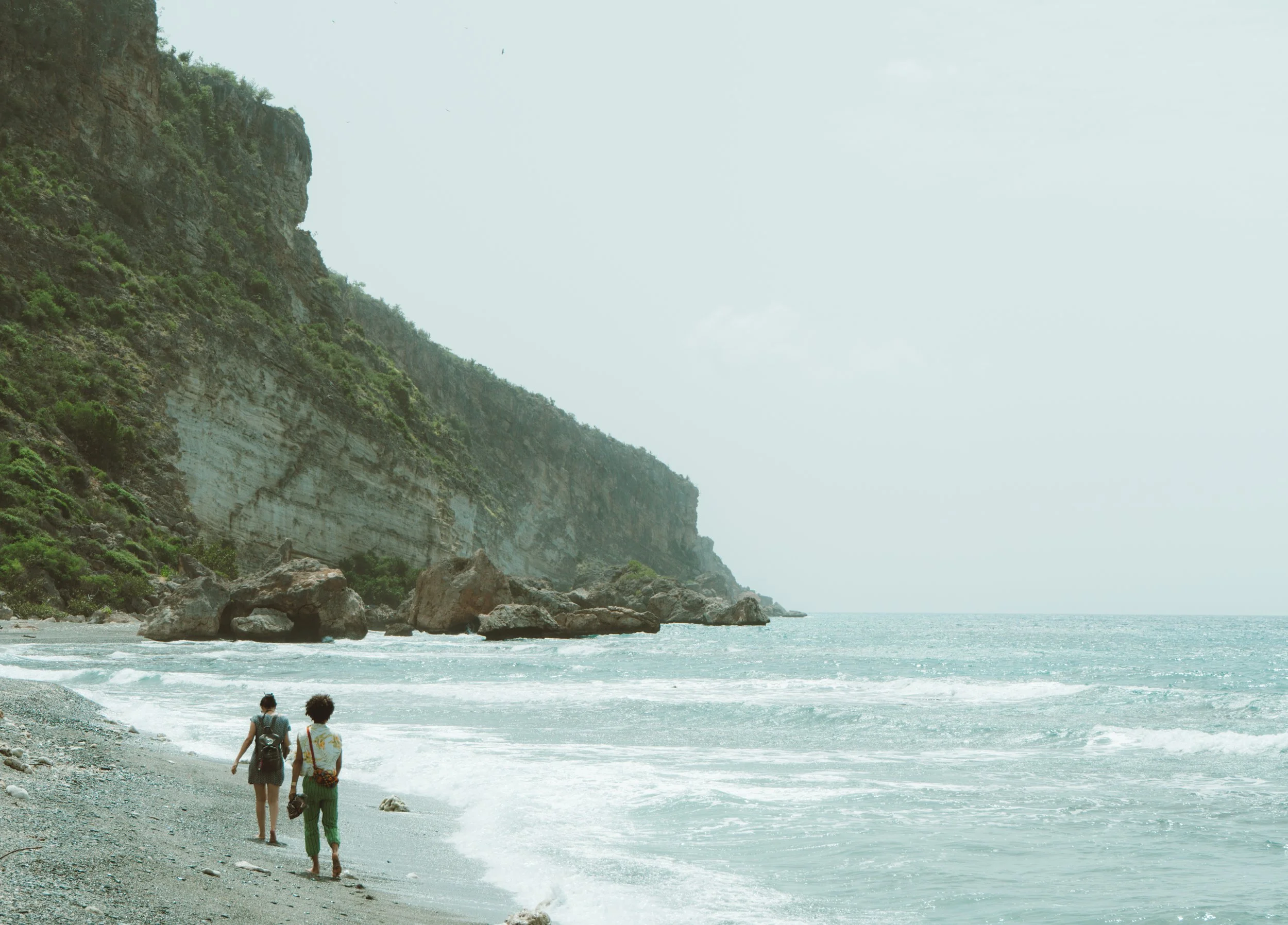 Two people walking along a rocky beach with cliffs covered in greenery on the left and the ocean on the right, under an overcast sky.