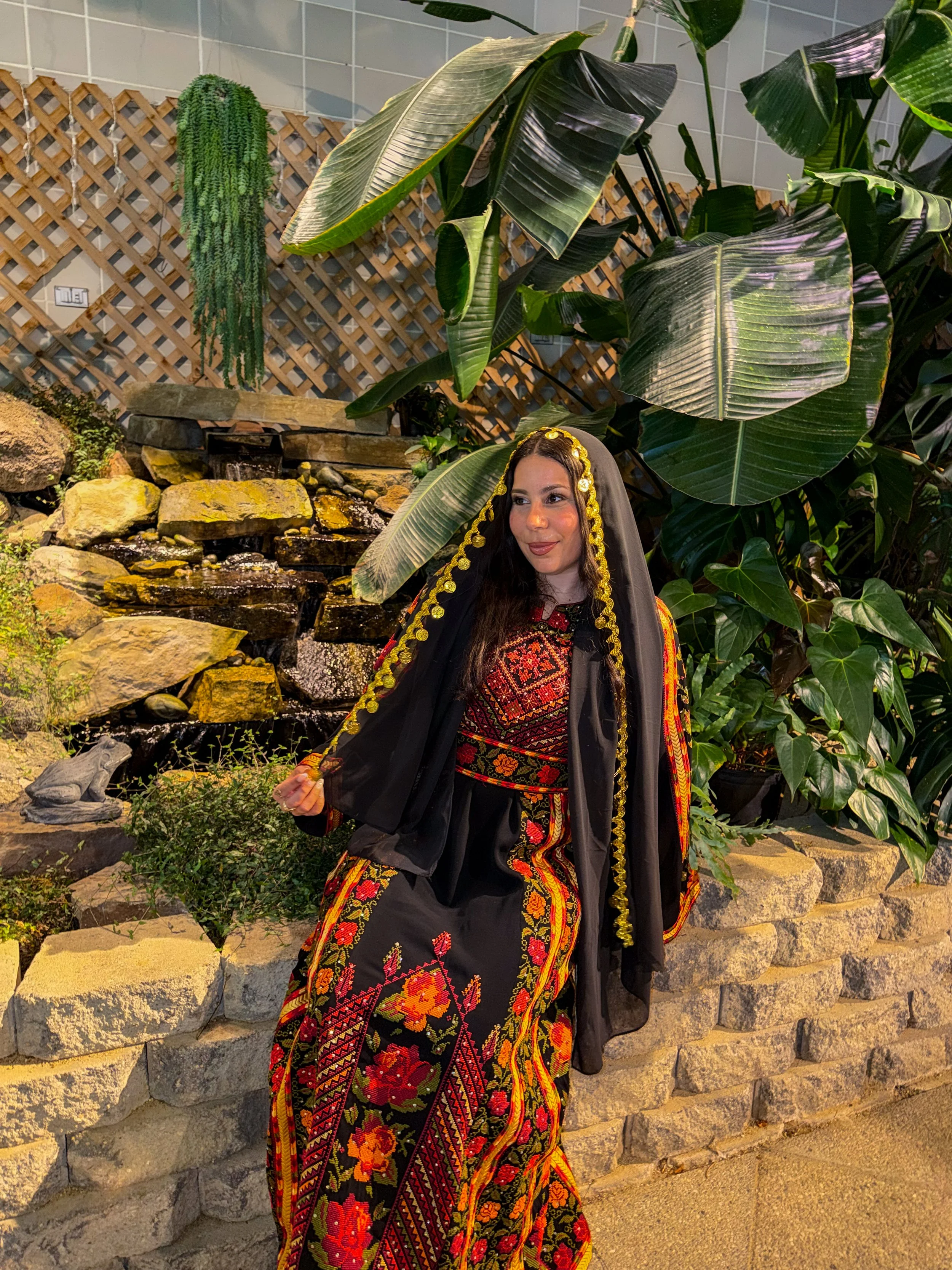 A woman wearing a black and red embroidered traditional dress and a black veil with gold trim, standing in front of large green tropical plants and a stone water feature with rocks and water.
