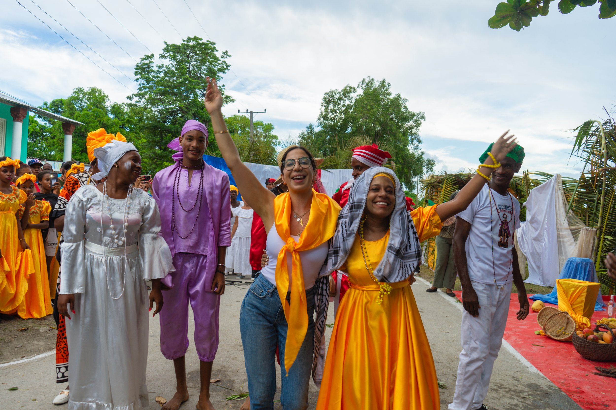 A diverse group of people dressed in traditional and colorful clothing participating in an outdoor cultural celebration or festival, smiling and waving.