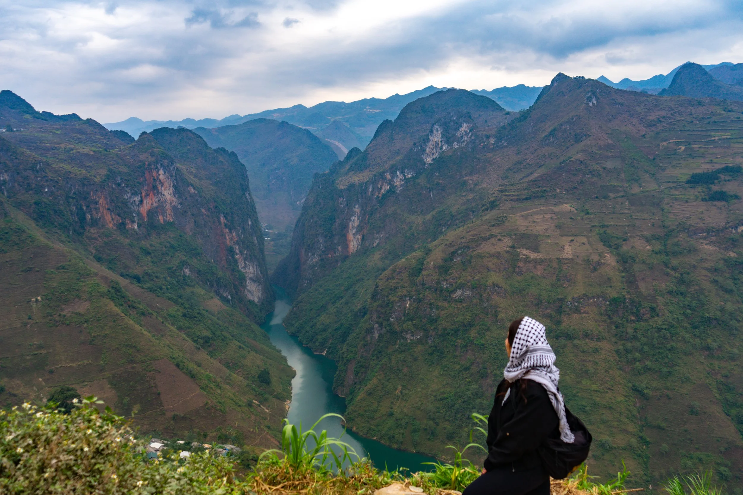 A woman wearing a black jacket and a checkered headscarf looks at a lush green mountainous landscape with steep hills and a river running through the valley below.