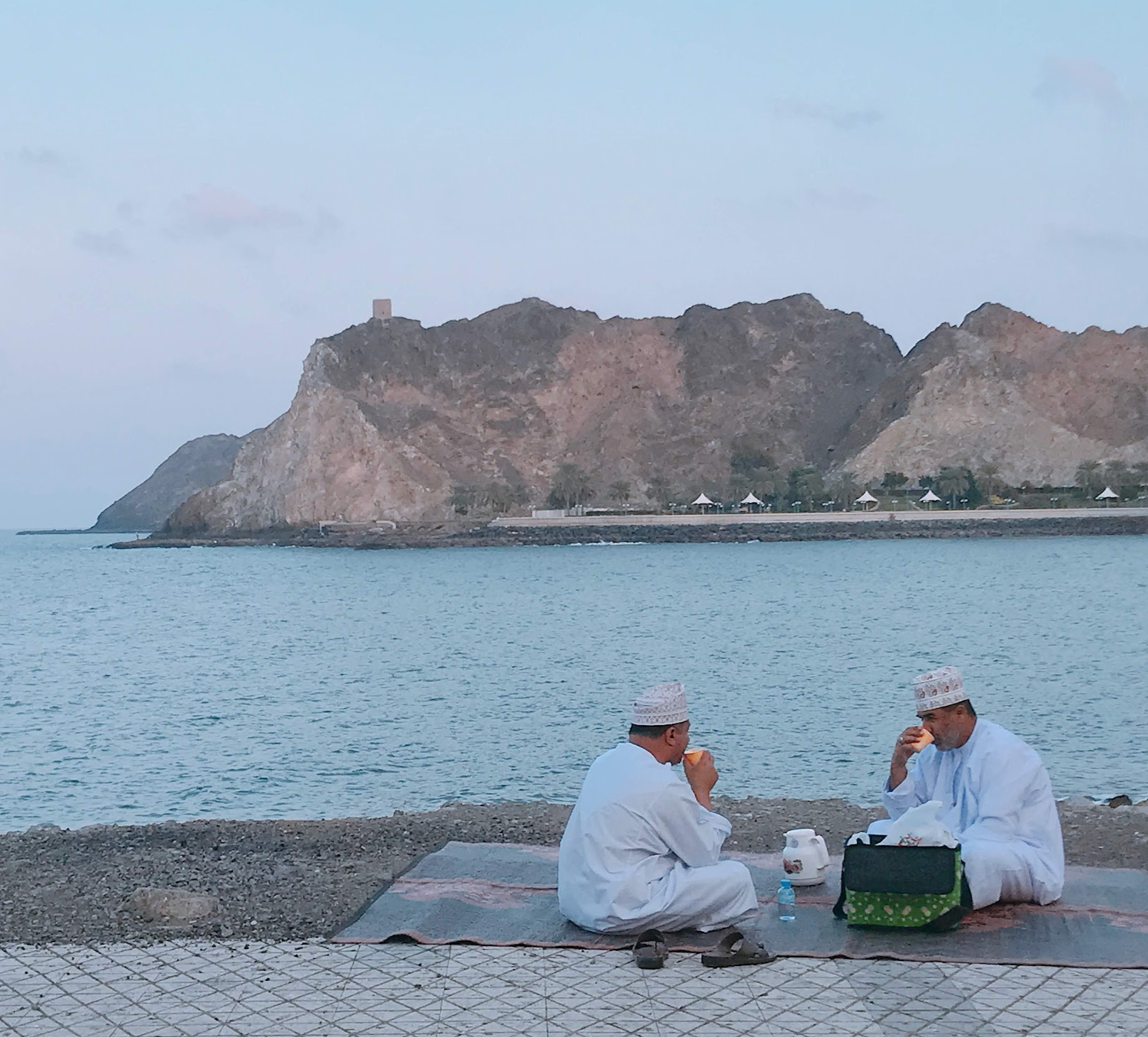 Two men in white Islamic clothing and hats sit on a mat outdoors near water, eating. In the background, a rocky hillside with a small tower and a row of white tents, with a cloudy sky.