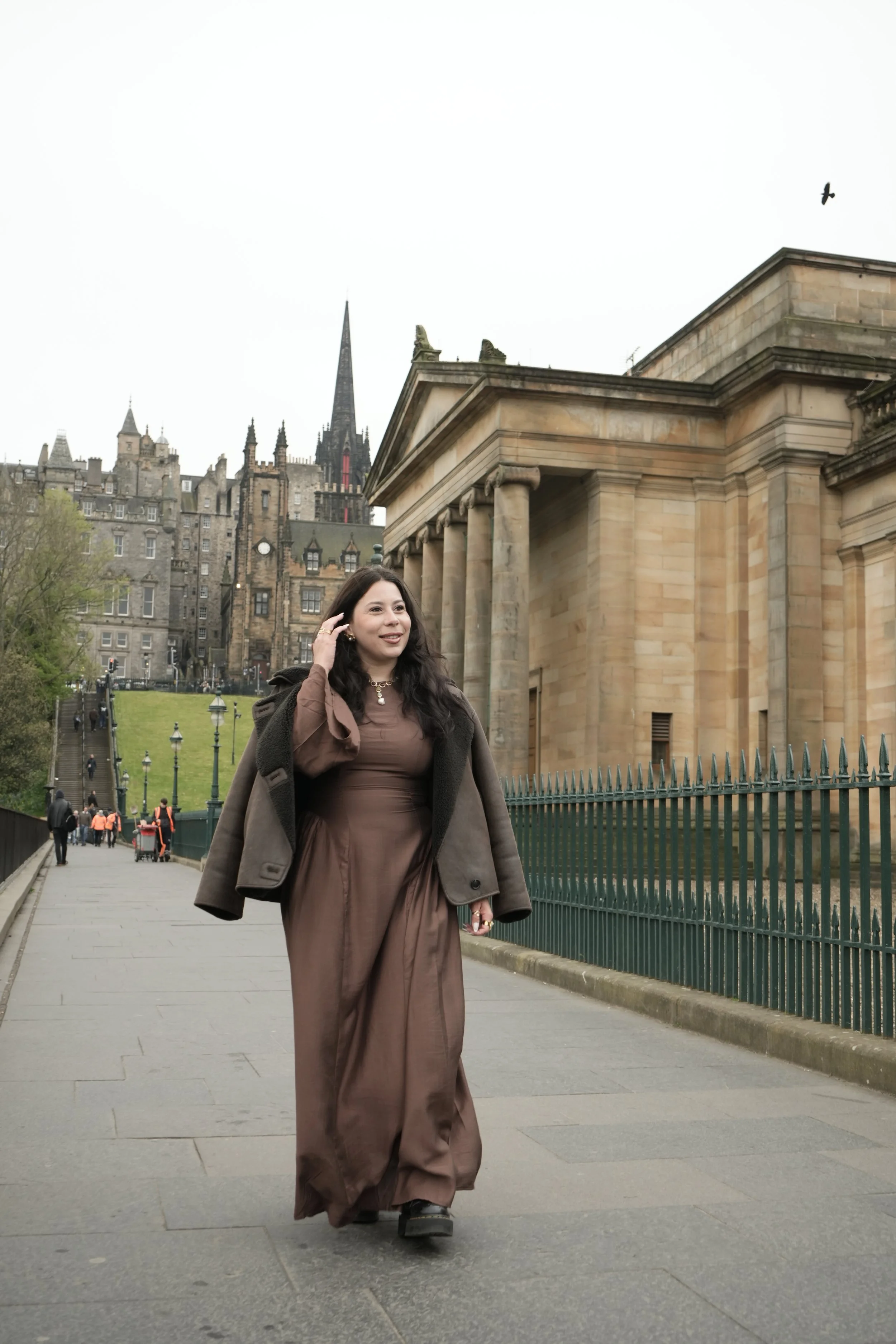 A woman wearing a brown gown and black boots walking on a paved street in front of historic buildings with gothic spires in Edinburgh, Scotland.