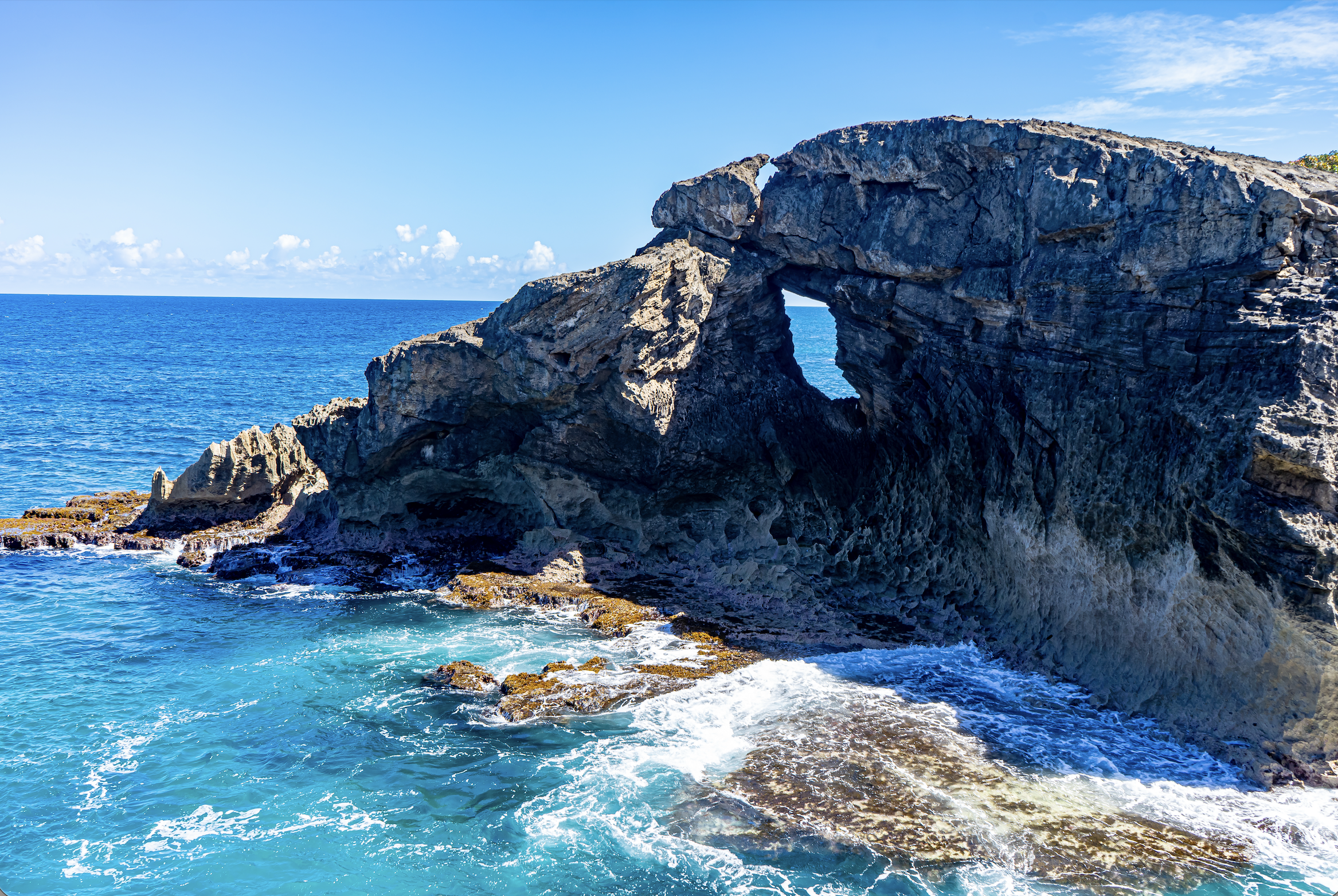 A large rock formation with a natural arch over the ocean, blue sky with some clouds in the background, and water breaking against the rocks at the base.