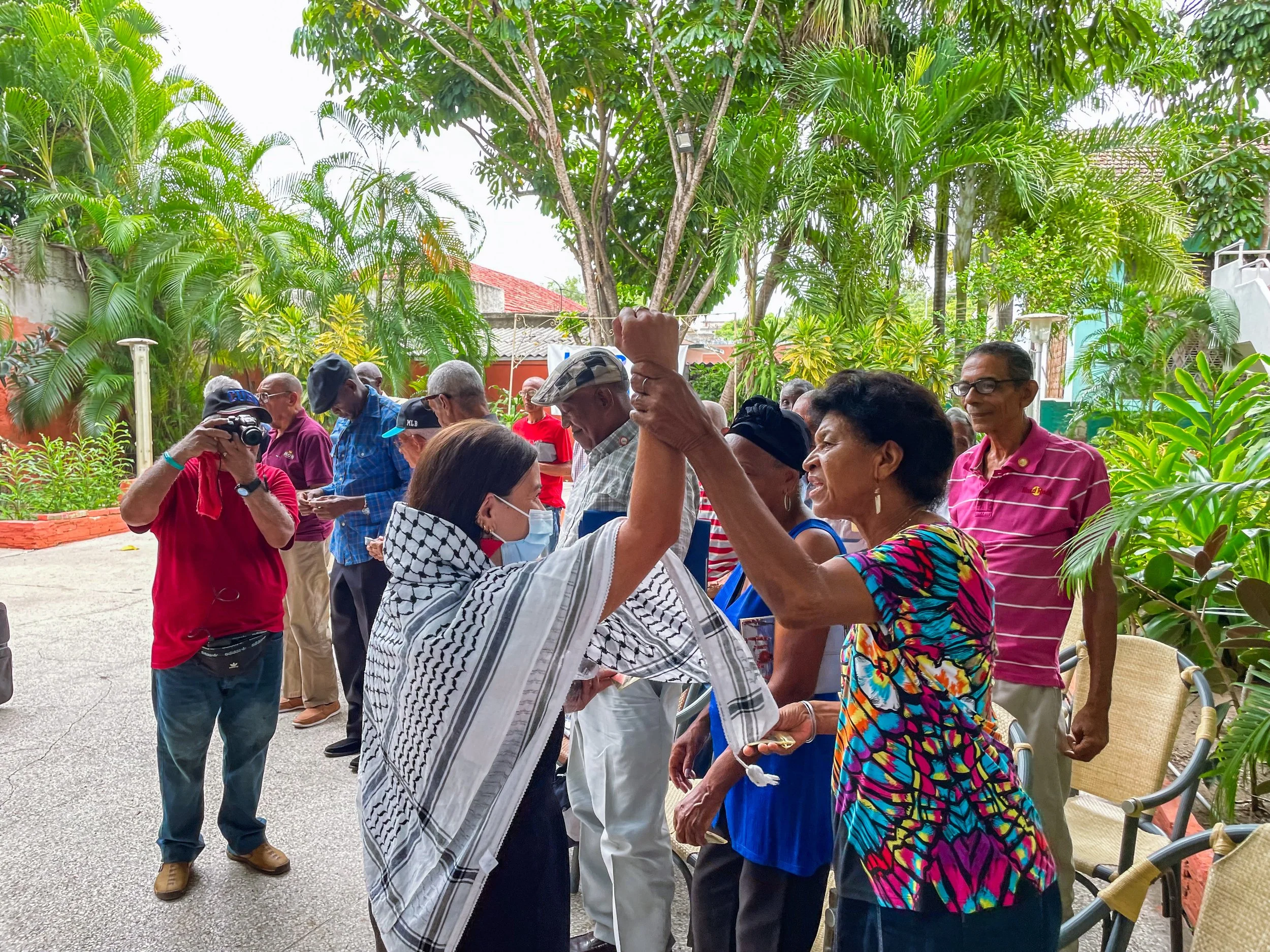 A group of diverse people, including men and women, gathered outdoors surrounded by lush green trees and plants. Some individuals are engaged in conversation, taking photos, or using their phones, with one person raising a hand and others smiling. They appear to be at a social or community event in a garden or backyard setting.