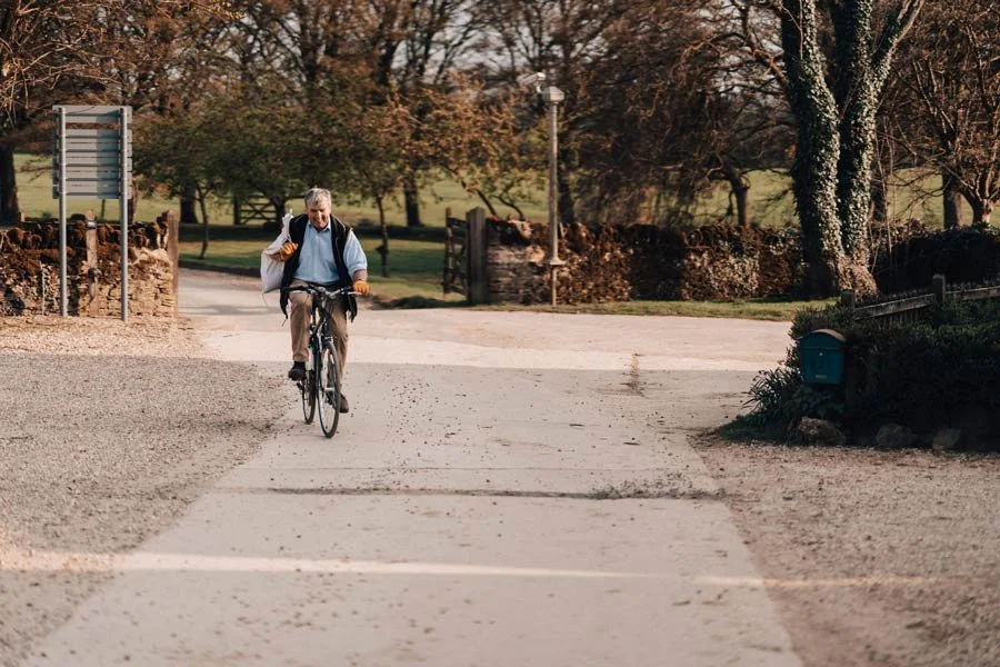 An older man riding a bicycle on a dirt path in a park during daytime, with trees and outdoor scenery around him.