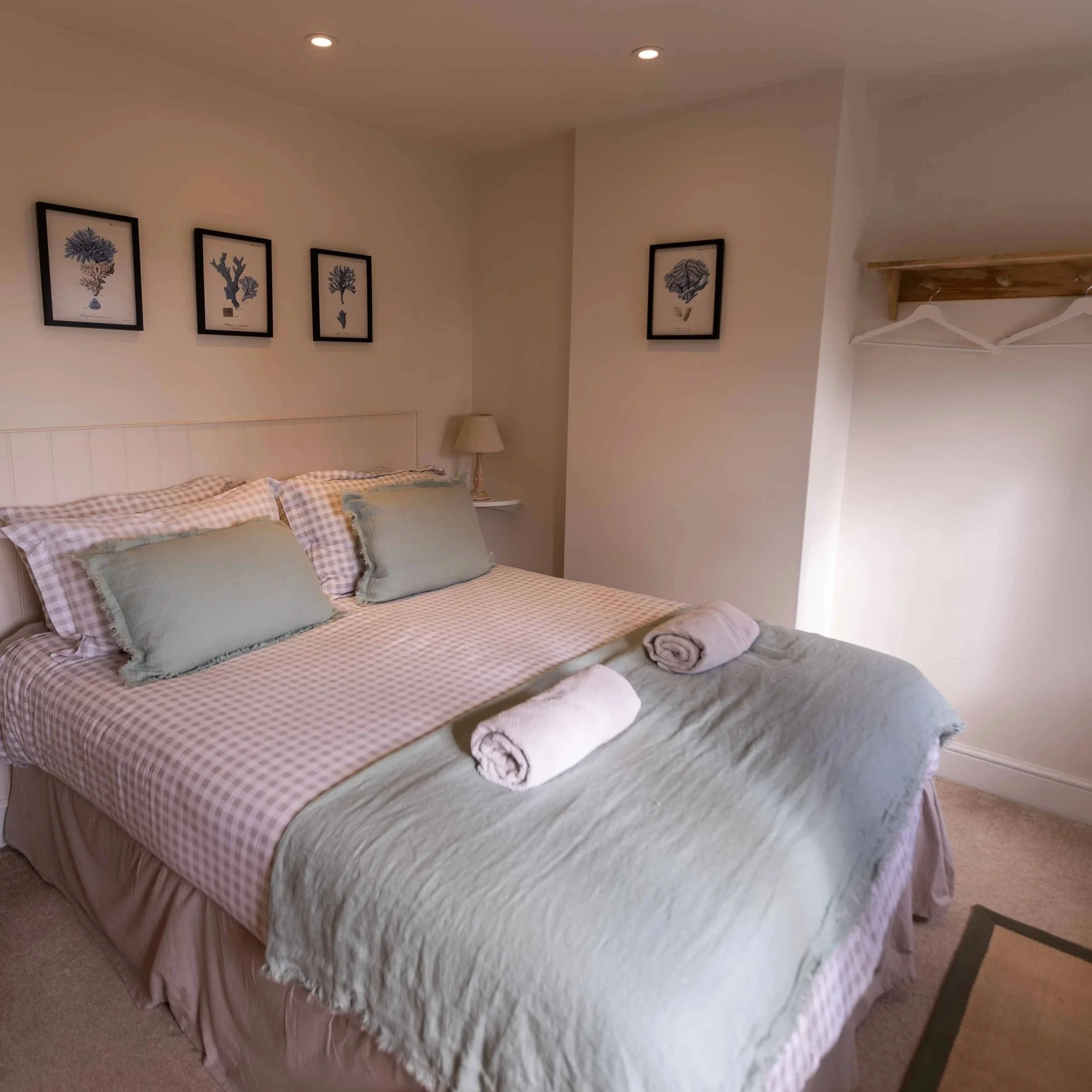 A neatly made bed with checkered and plain pillows, topped with rolled towels, in a cozy bedroom with framed coral shell artwork on the wall, a small lamp on a bedside table, and a wall shelf with empty hangers.