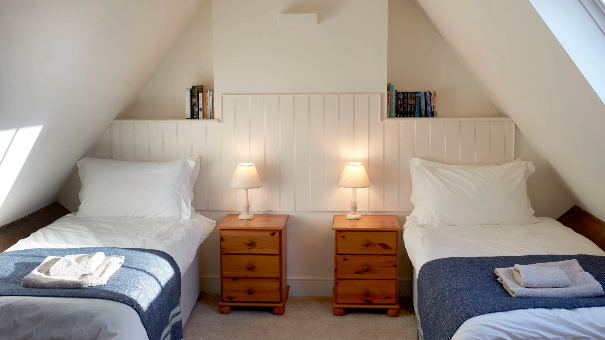 A cozy bedroom with two single beds featuring white bedding and blue blankets. There are two wooden nightstands with lamps between the beds. Each bed has a towel and folded clothes on it. The walls are painted white with a simple headboard design and