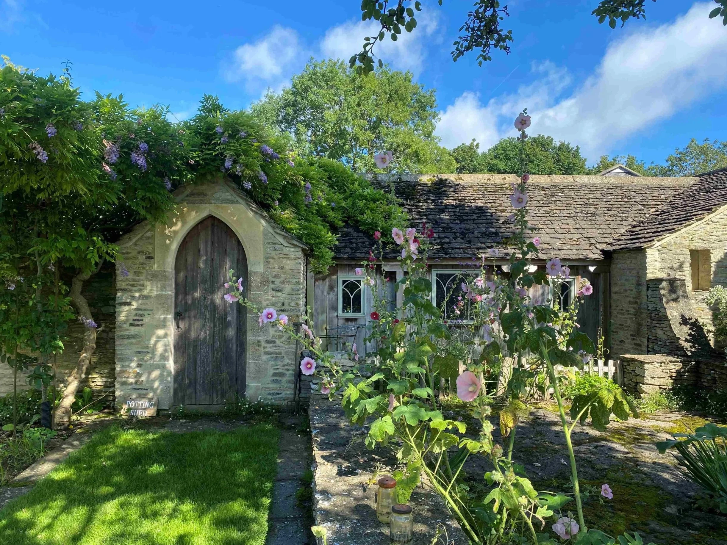 An old stone cottage with a rustic wooden door, surrounded by lush green trees and flowering plants, under a bright blue sky with some clouds.