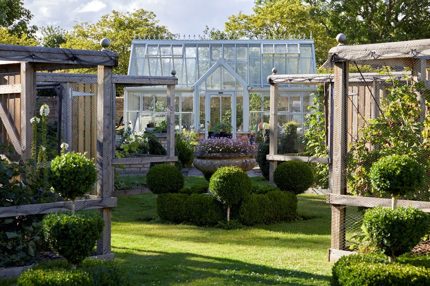 A well-maintained garden with a central stone planter filled with pink flowers, surrounded by neatly trimmed bushes and a lush green lawn. In the background, there are two glass greenhouses with metal frames and wooden garden structures with wire mes