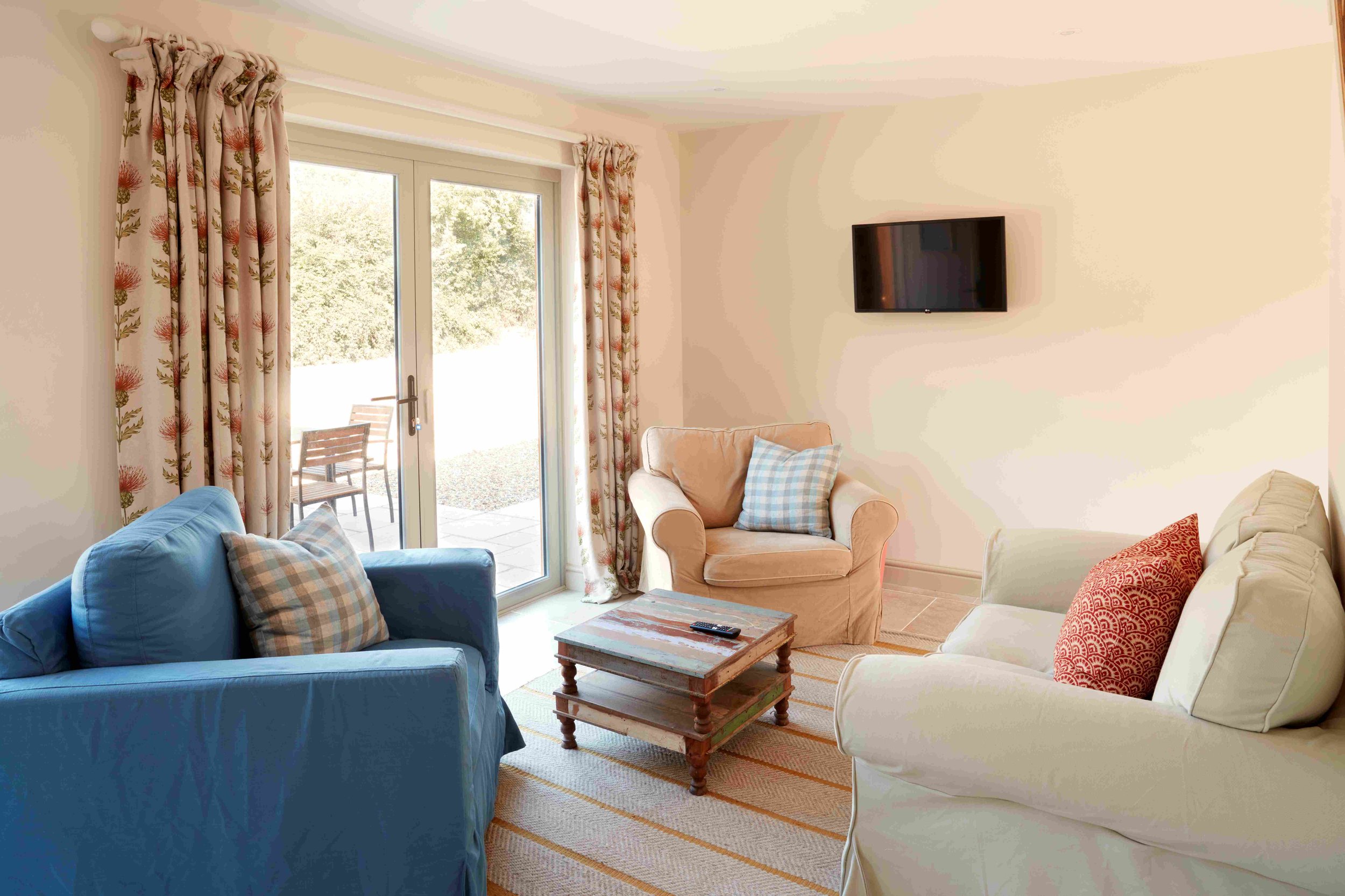 Living room with beige walls, a sliding glass door with floral curtains, two sofas (one blue with plaid pillows and one beige with red pillow), a wooden coffee table with a remote, a wall-mounted TV, and an outdoor patio with chairs visible through t