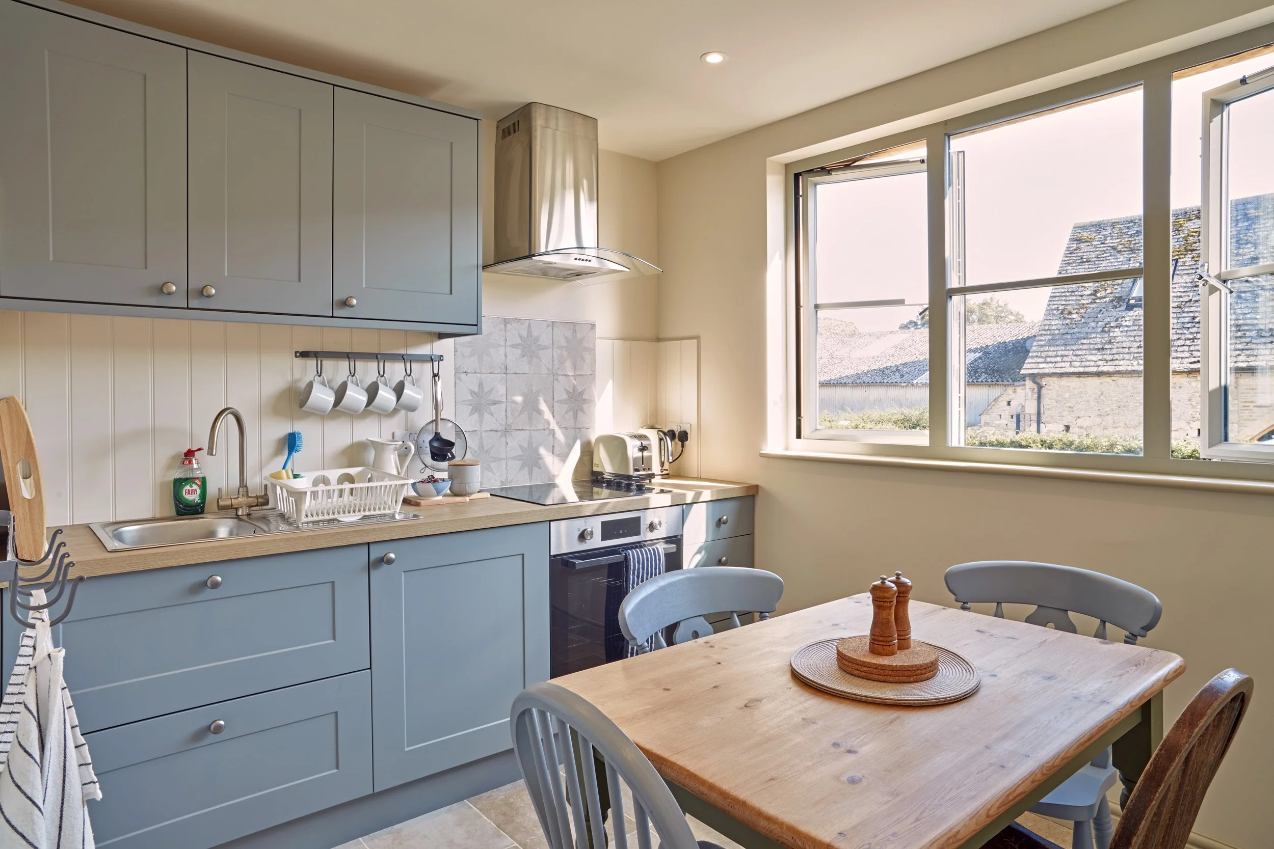 Kitchen with blue cabinets, stainless steel range hood, and a large window with a view of neighboring rooftops. There is a wooden dining table with chairs, a dish rack, and some kitchen appliances on the counter.
