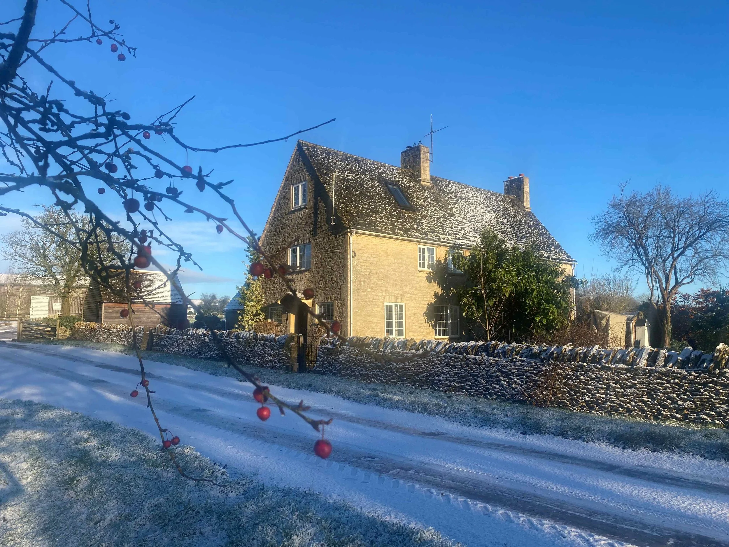 A snow-covered landscape featuring a large, stone house with a steep roof, surrounded by leafless trees and a stone wall under a clear blue sky.