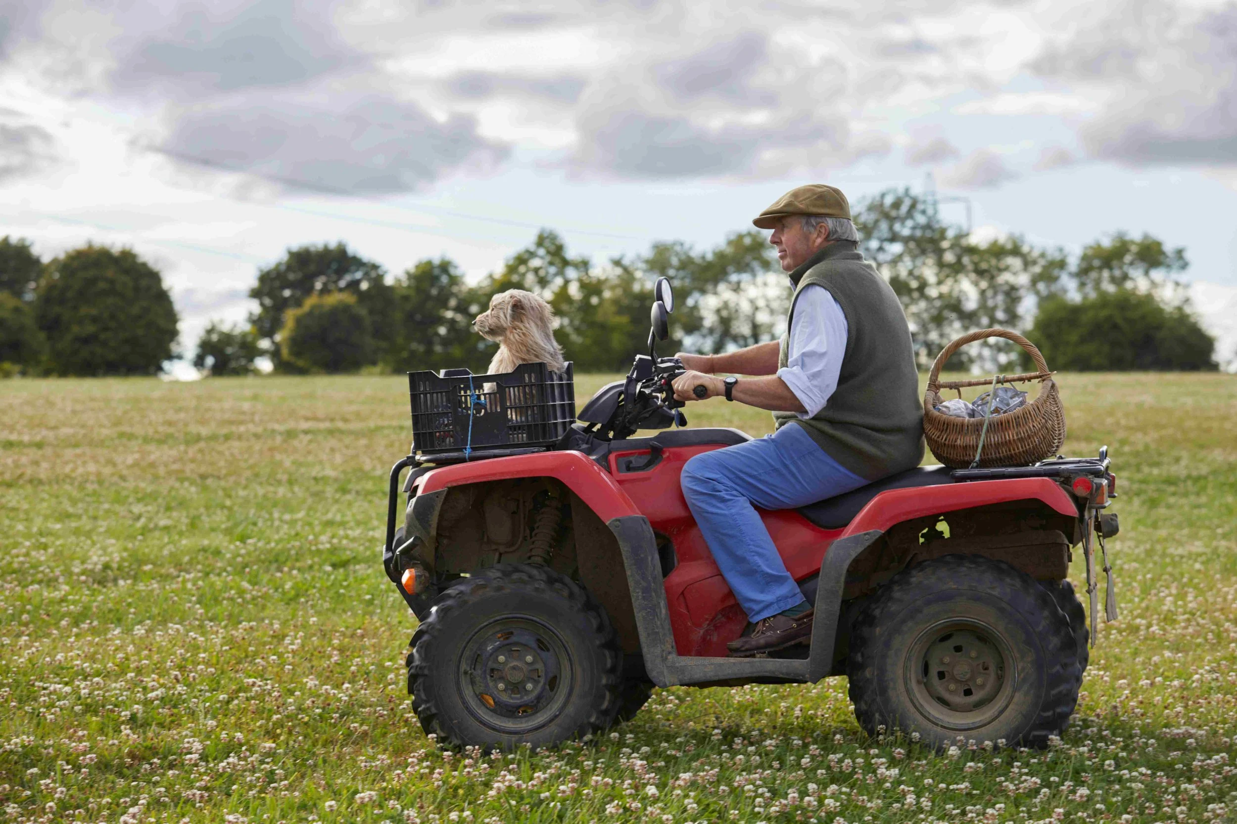 An elderly man riding a red all-terrain vehicle with a dog sitting in a basket in front, outdoors in a grassy field with trees and cloudy sky in the background.