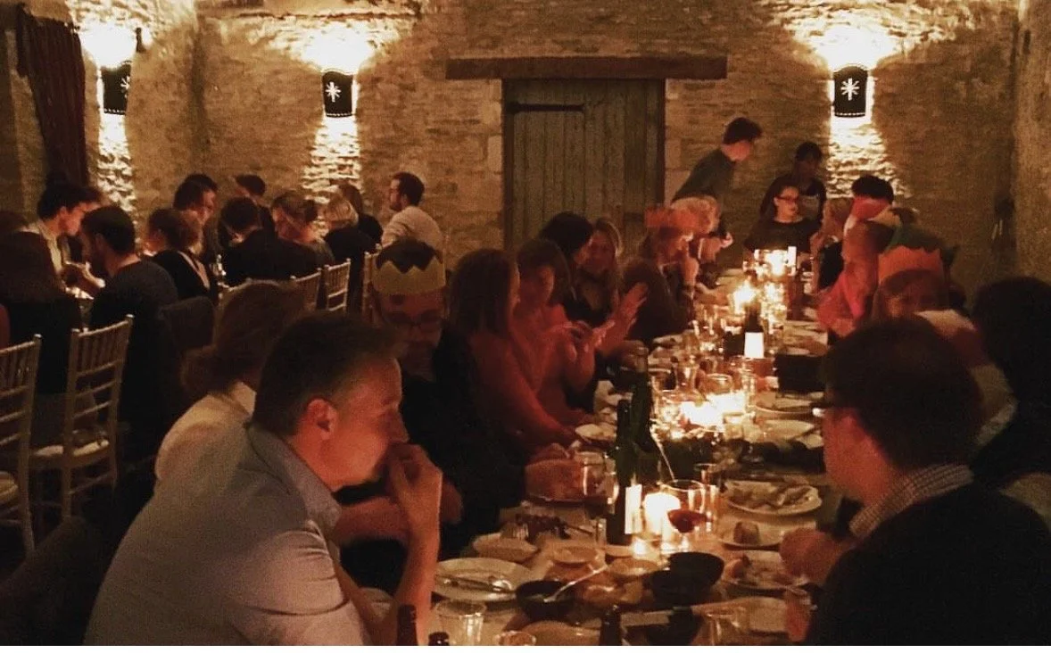 A long dining table with many people seated, enjoying a dinner in a dimly lit rustic stone-walled room with candlelights.