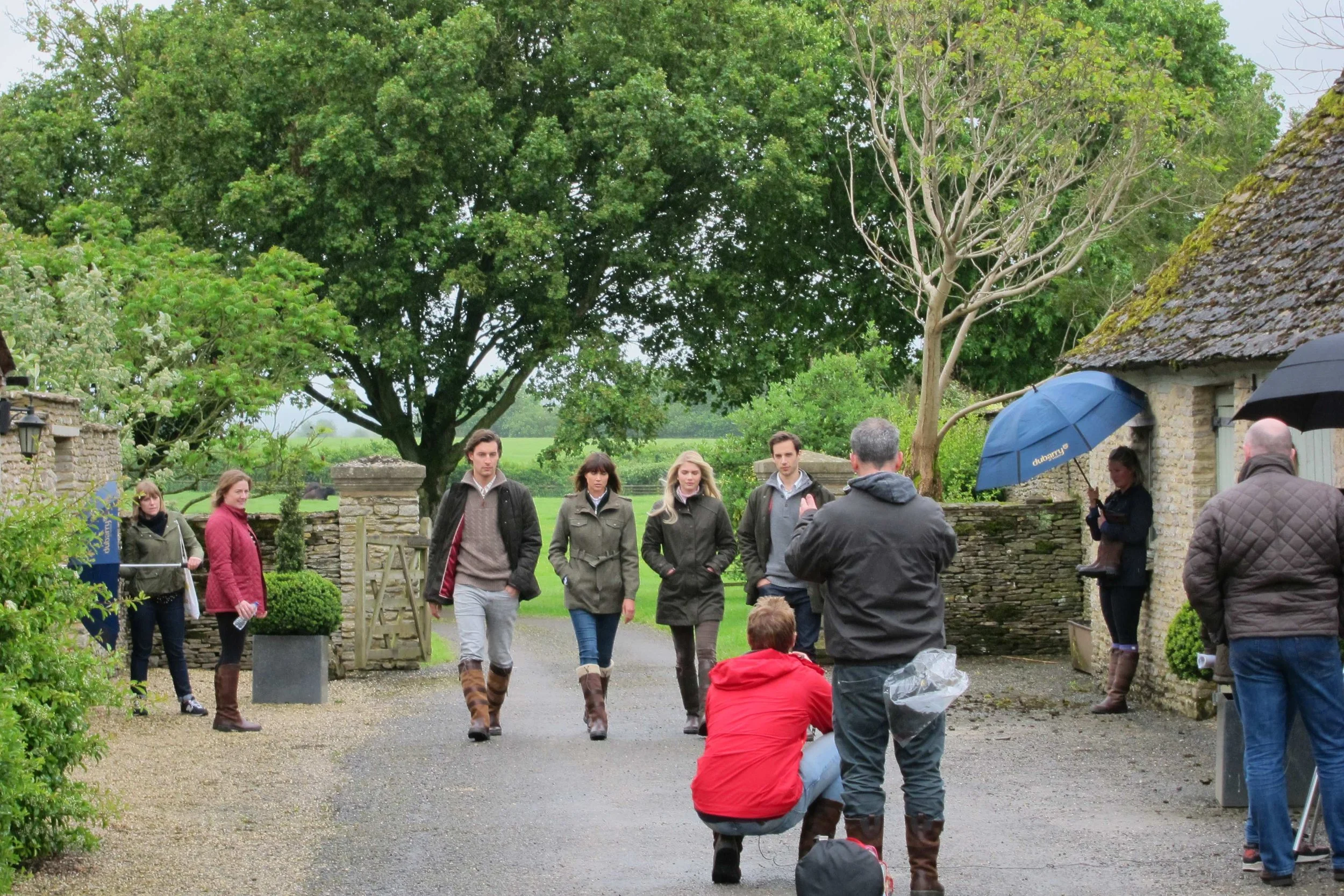 Group of people gathered outdoors in front of a stone building, with large trees and greenery in the background, some holding umbrellas and others standing or crouching.