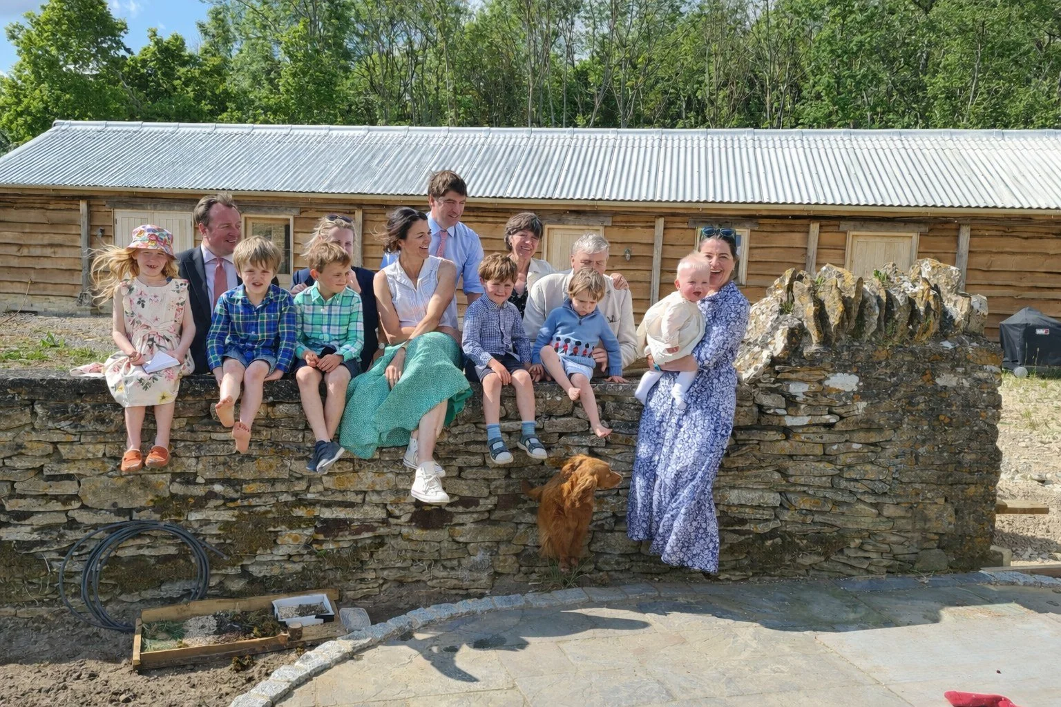 Group of people, including children and adults, sitting and standing on a stone wall outdoors during daytime. Some are smiling and interacting, with a woman holding a small child, and there is a dog below the wall. A rustic wooden building with a metal roof is in the background, surrounded by green trees.