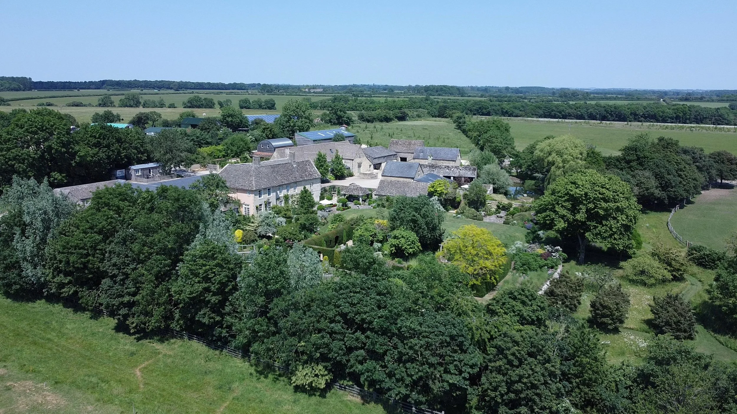 A rural landscape with green trees, fields, and a cluster of old stone buildings with tiled roofs, surrounded by lush gardens and open land under a clear blue sky.