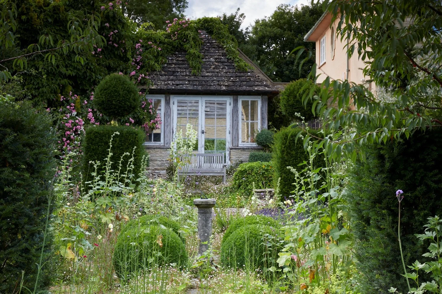 A lush garden with various green shrubs, flowering plants, and tall herbs leading to a cozy, rustic house with a bay window and a small front porch with a bench. The scene is surrounded by tall trees and a neighboring house is visible on the right.