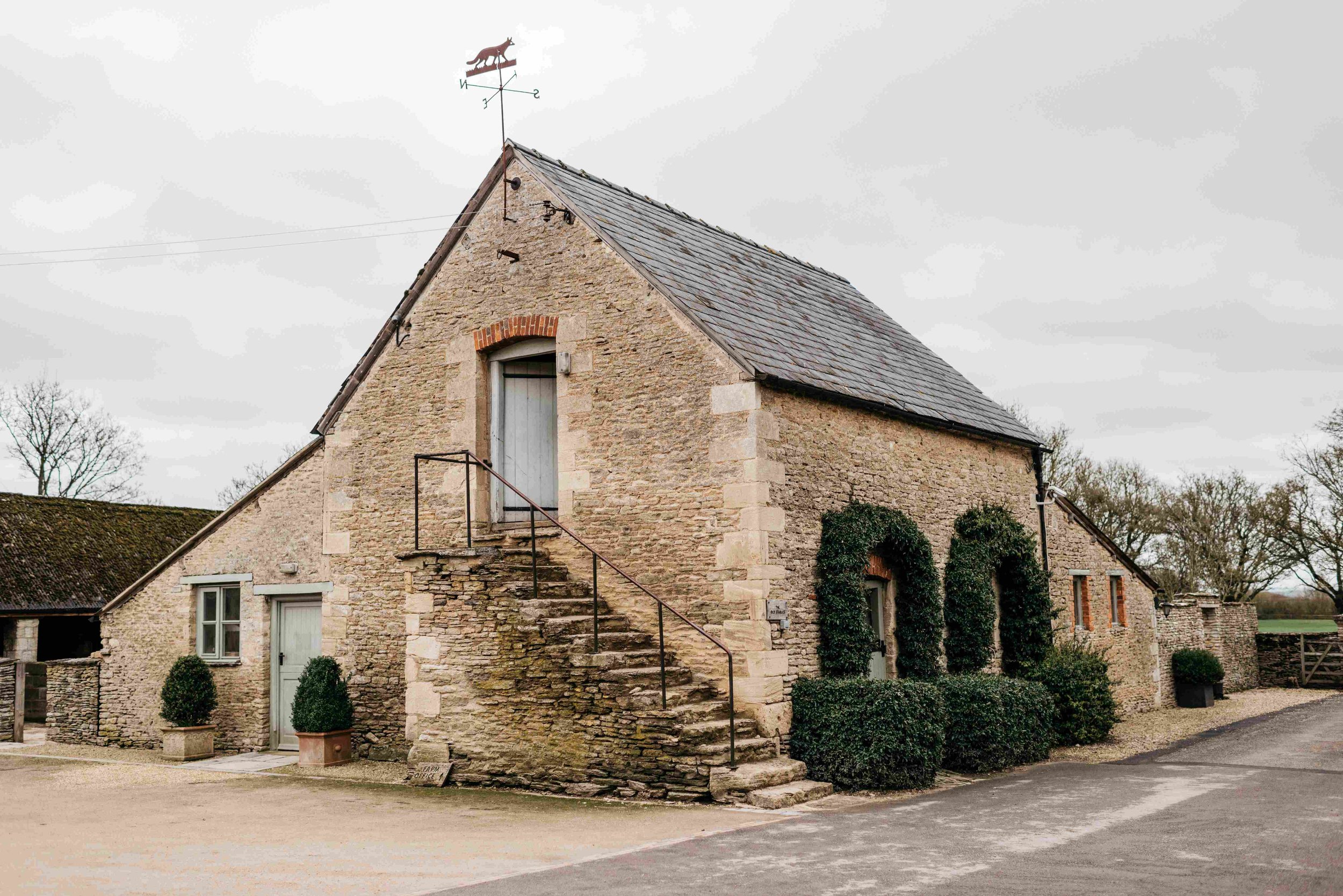 A stone building with a steep roof, small windows, and a staircase leading to a door with a sign that says 'Farm Office.' The building has climbing ivy and is set in a rural area with trees and a gravel road.