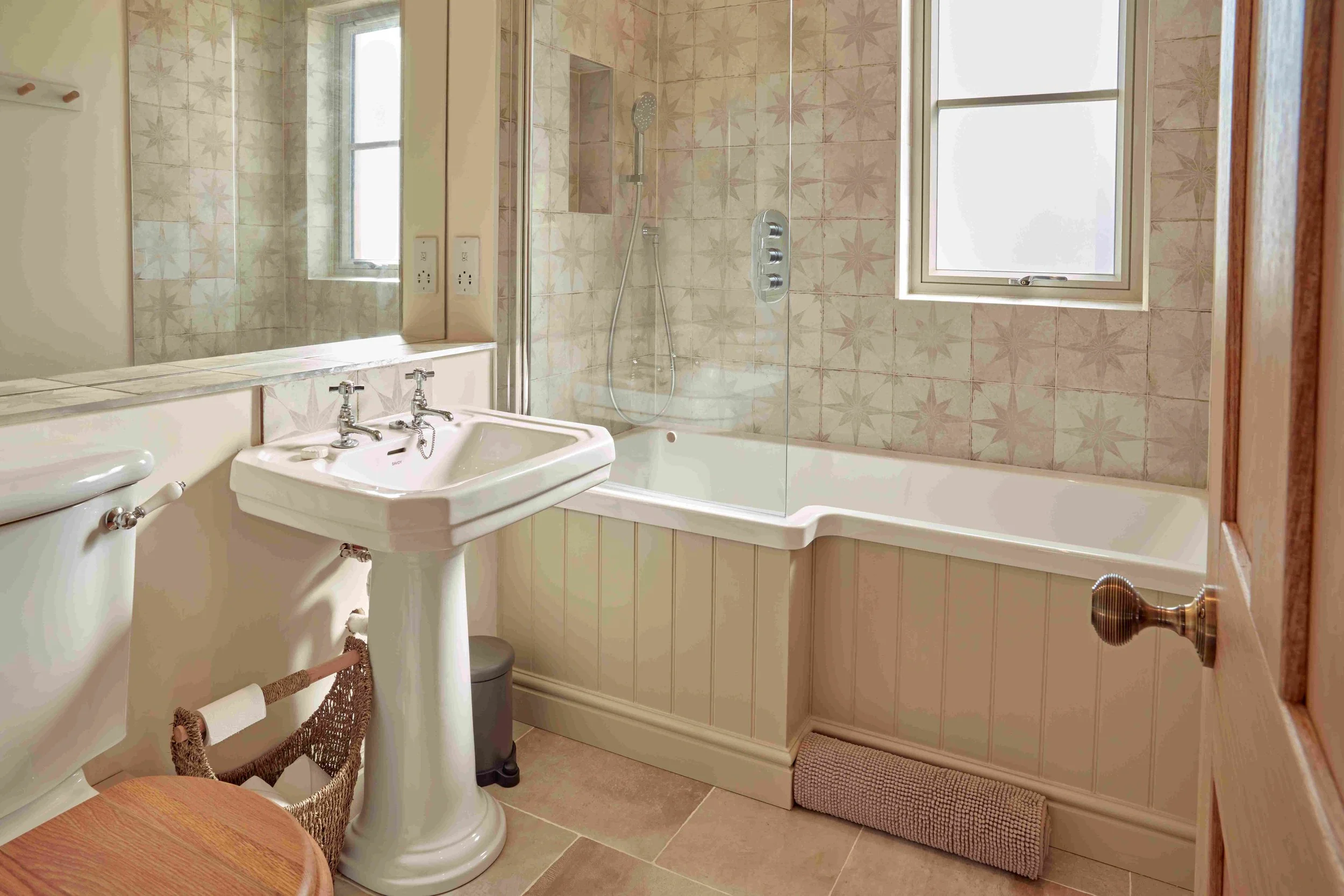 A small vintage-style bathroom with a pedestal sink, toilet, and a bathtub with a clear glass shower door. The walls are tiled with beige star-patterned tiles. There is a window above the bathtub, a mirror on the wall, and a woven basket under the si