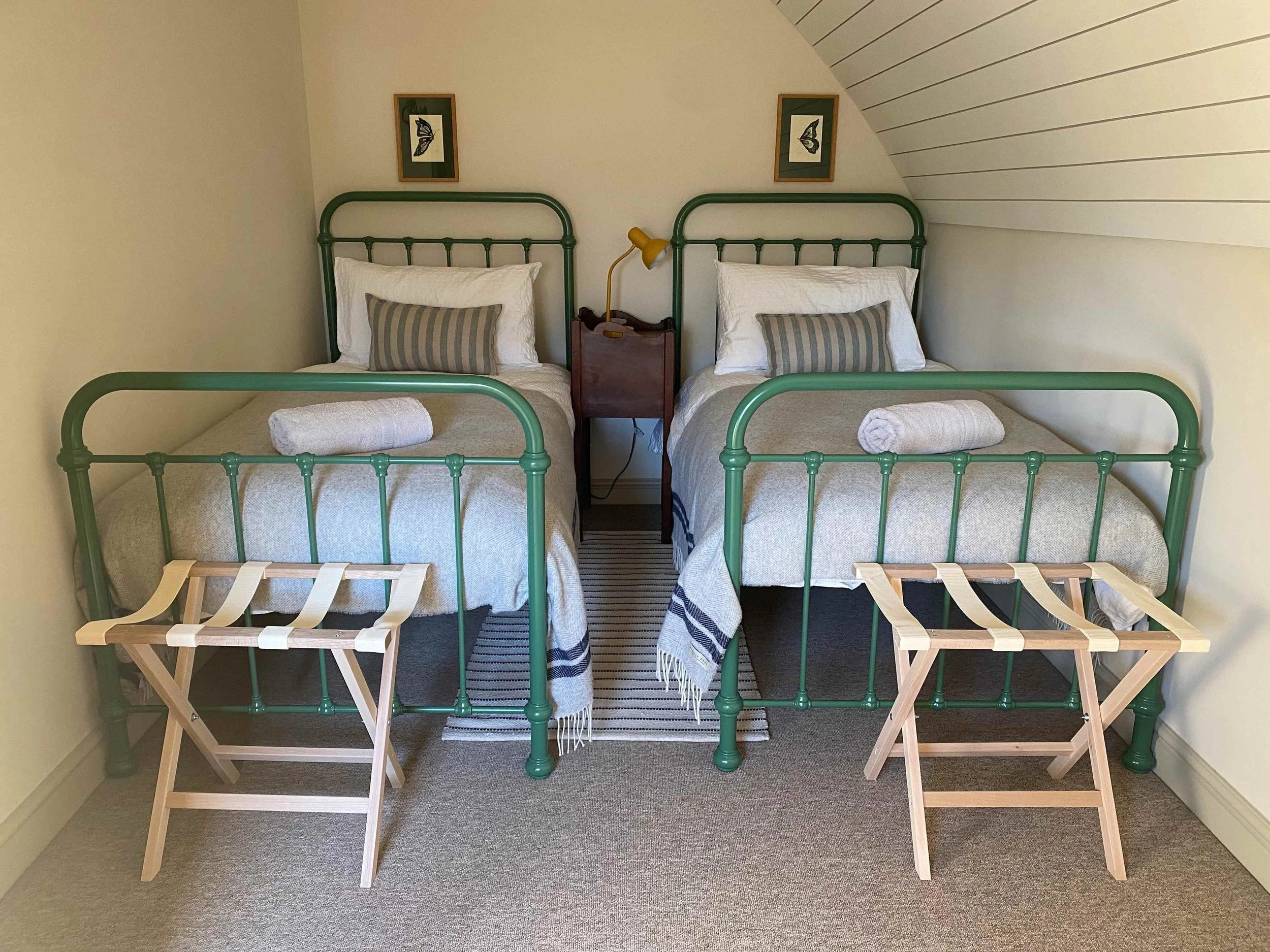 Two single beds with green metal frames in a small room, each with a pillow, beige bedspread, and a towel. There are wooden luggage racks in front of the beds, a small wooden nightstand with a yellow desk lamp, and framed butterfly artwork above each
