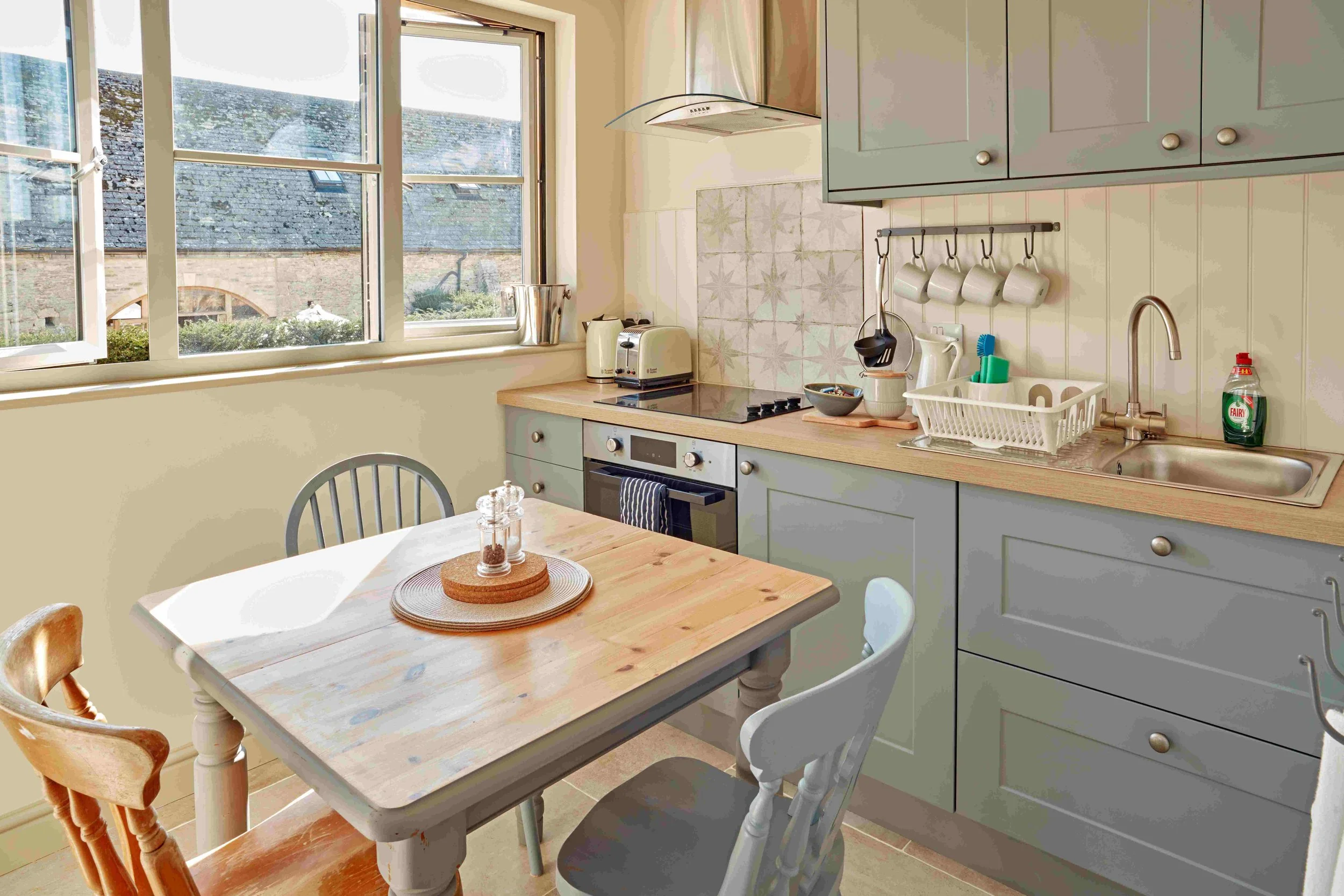 A cozy kitchen with a wooden table and several chairs near a large window, teal cabinets, a stainless steel sink, a stove, and various kitchen appliances.
