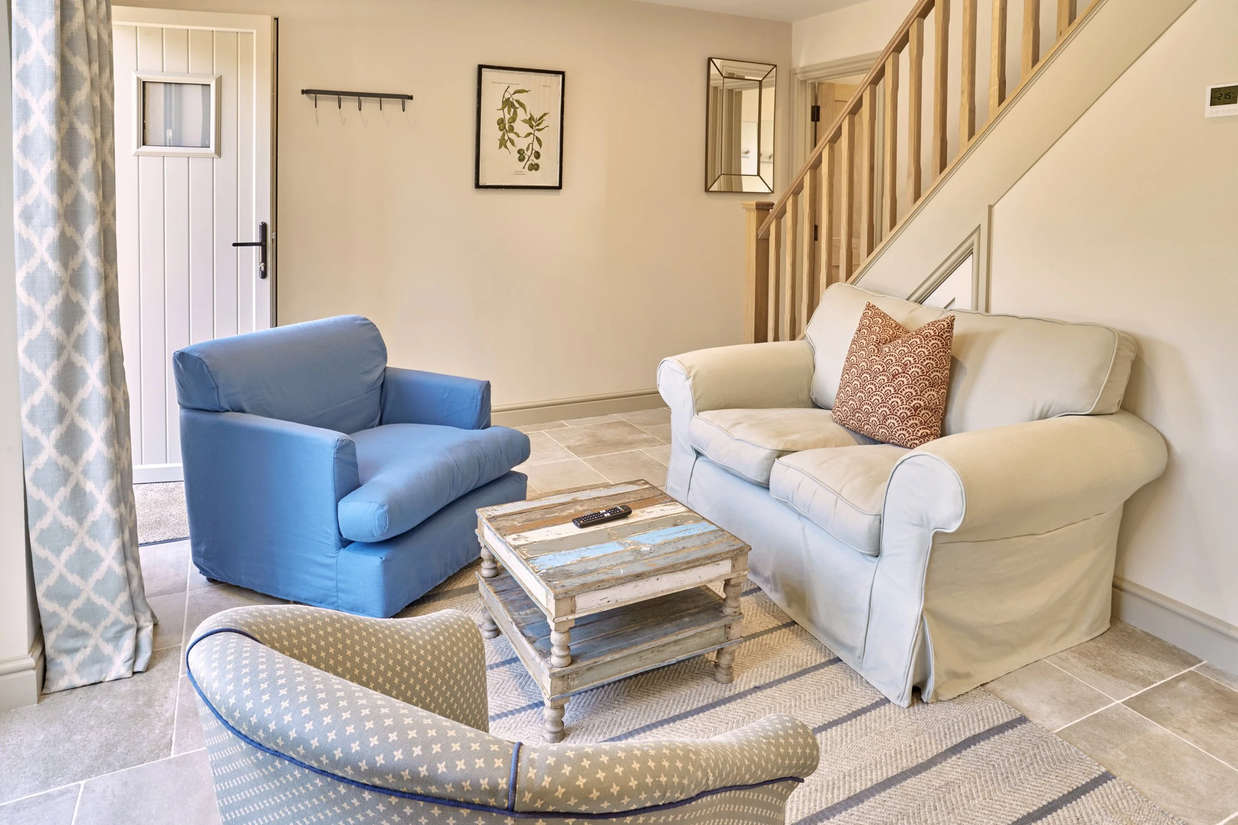 A living room with a beige sofa, a blue armchair, a pair of patterned chairs, and a rustic coffee table. The background includes a staircase with wooden railing, a wall with framed art and a mirror, a white door with a small window, and a digital the