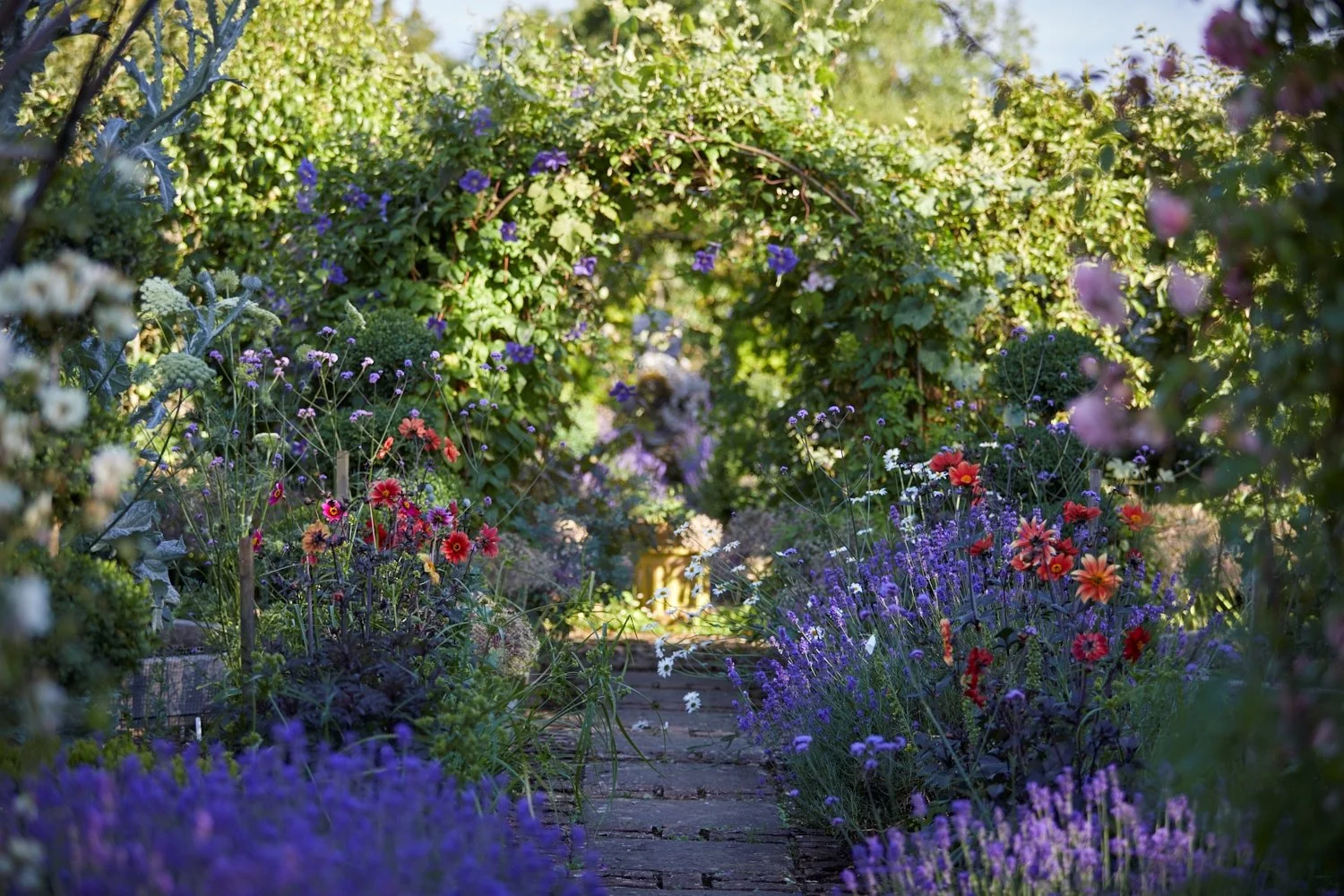 A lush garden pathway surrounded by colorful flowers and greenery, with sunlight filtering through the plants.