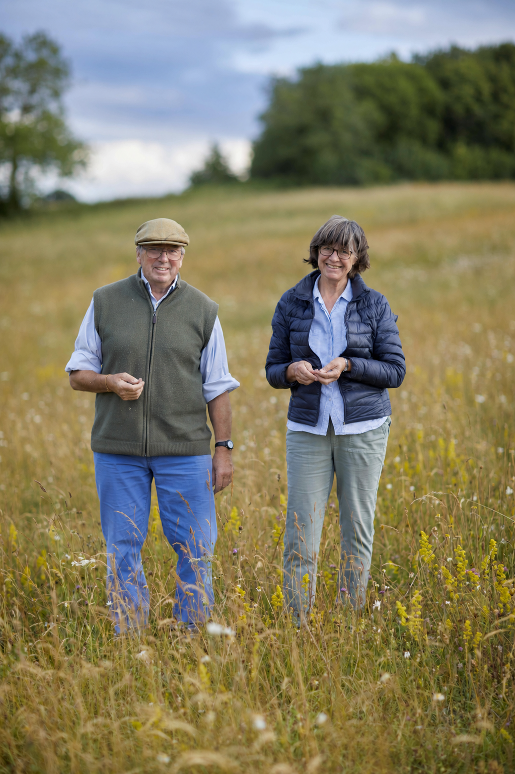 A man and woman standing in a grassy field, smiling, with trees and a cloudy sky in the background.