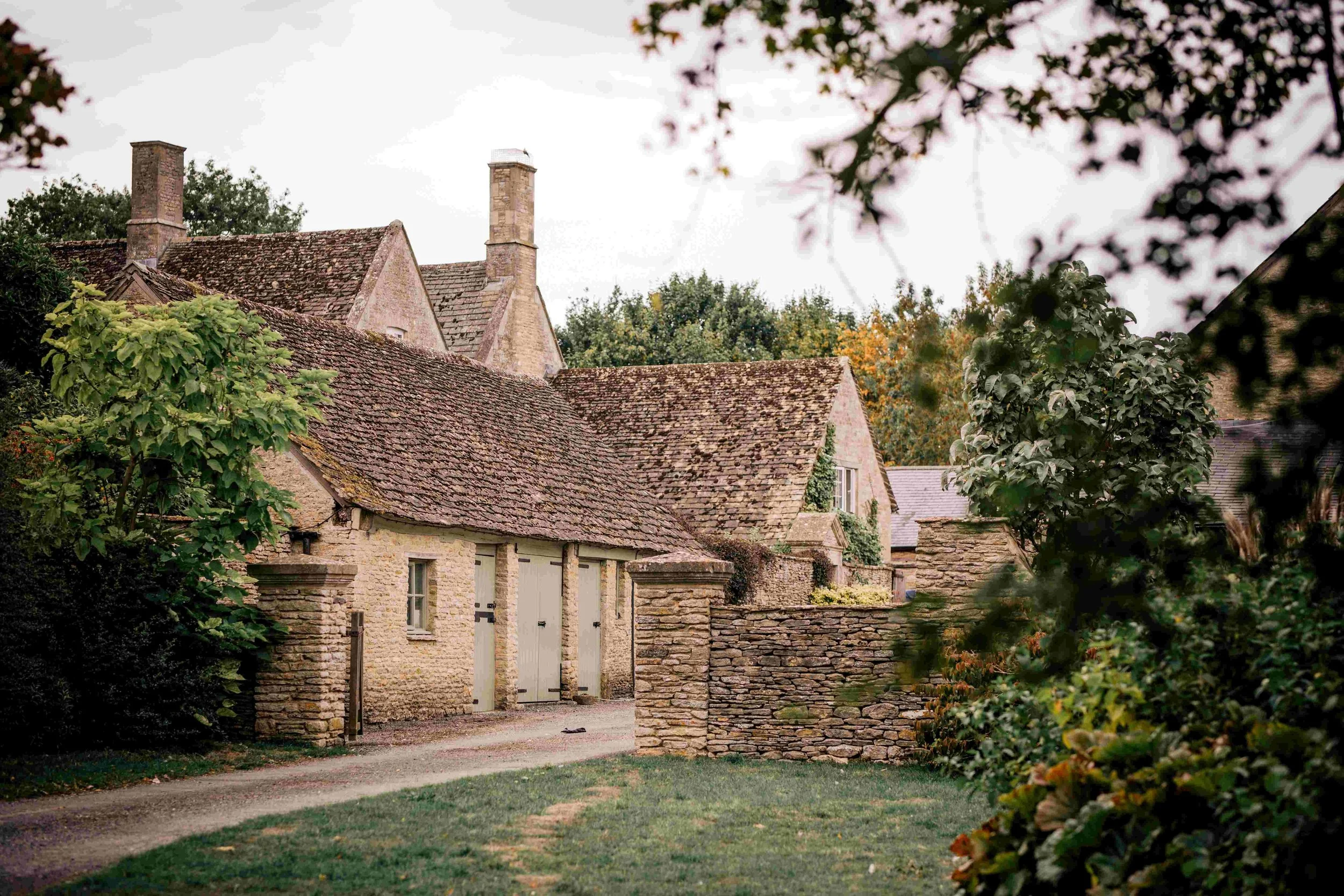 View of a stone cottage with a tiled roof, surrounded by trees and greenery, framed by branches and leaves in the foreground.