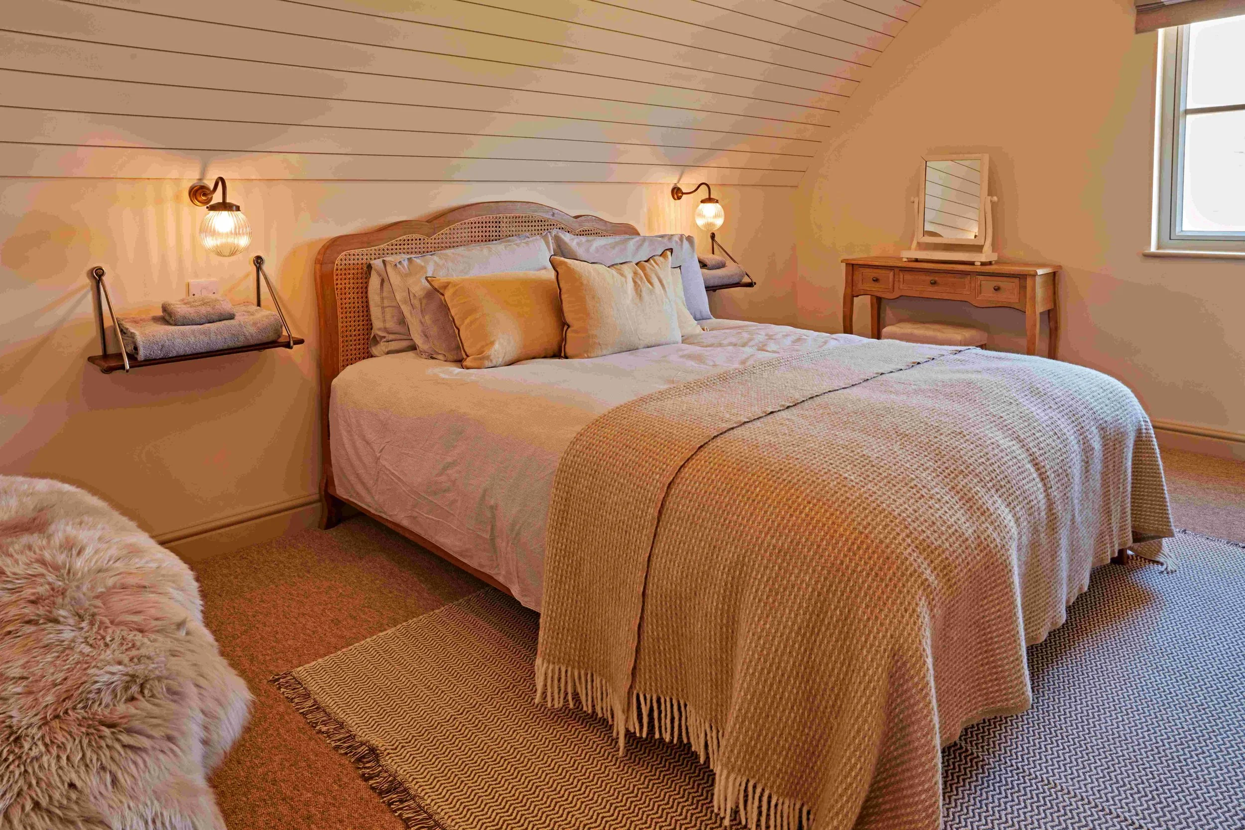 A cozy bedroom with a bed covered in beige and pastel-colored pillows, a wooden headboard, and matching bedside lamps with round glass shades mounted on the wall. There's a small wooden vanity with a mirror and a window providing natural light. The s