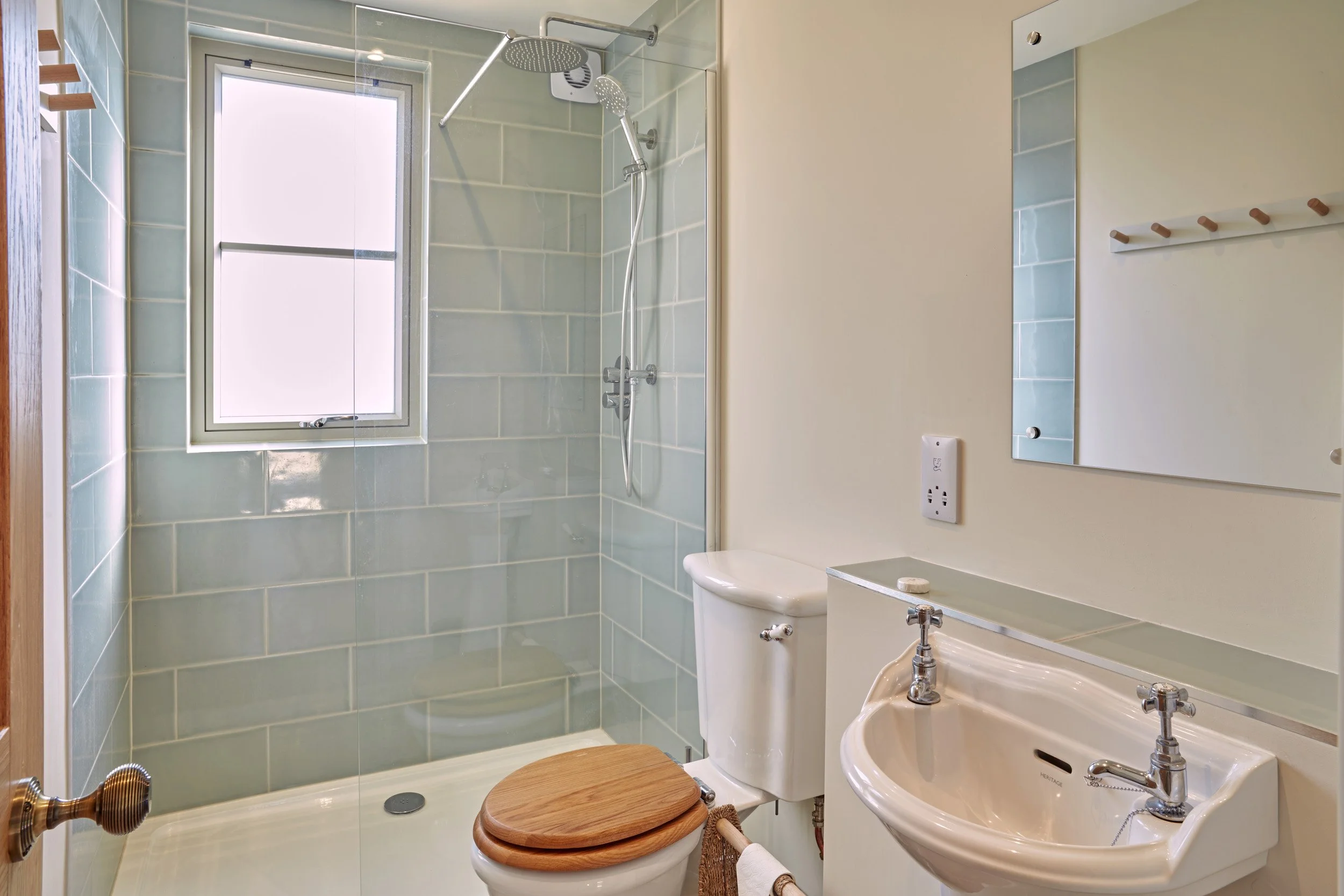 Small bathroom with a shower area enclosed with a glass door, a frosted window, a toilet with a wooden seat, and a small white sink with dual taps, mirror, and beige walls.
