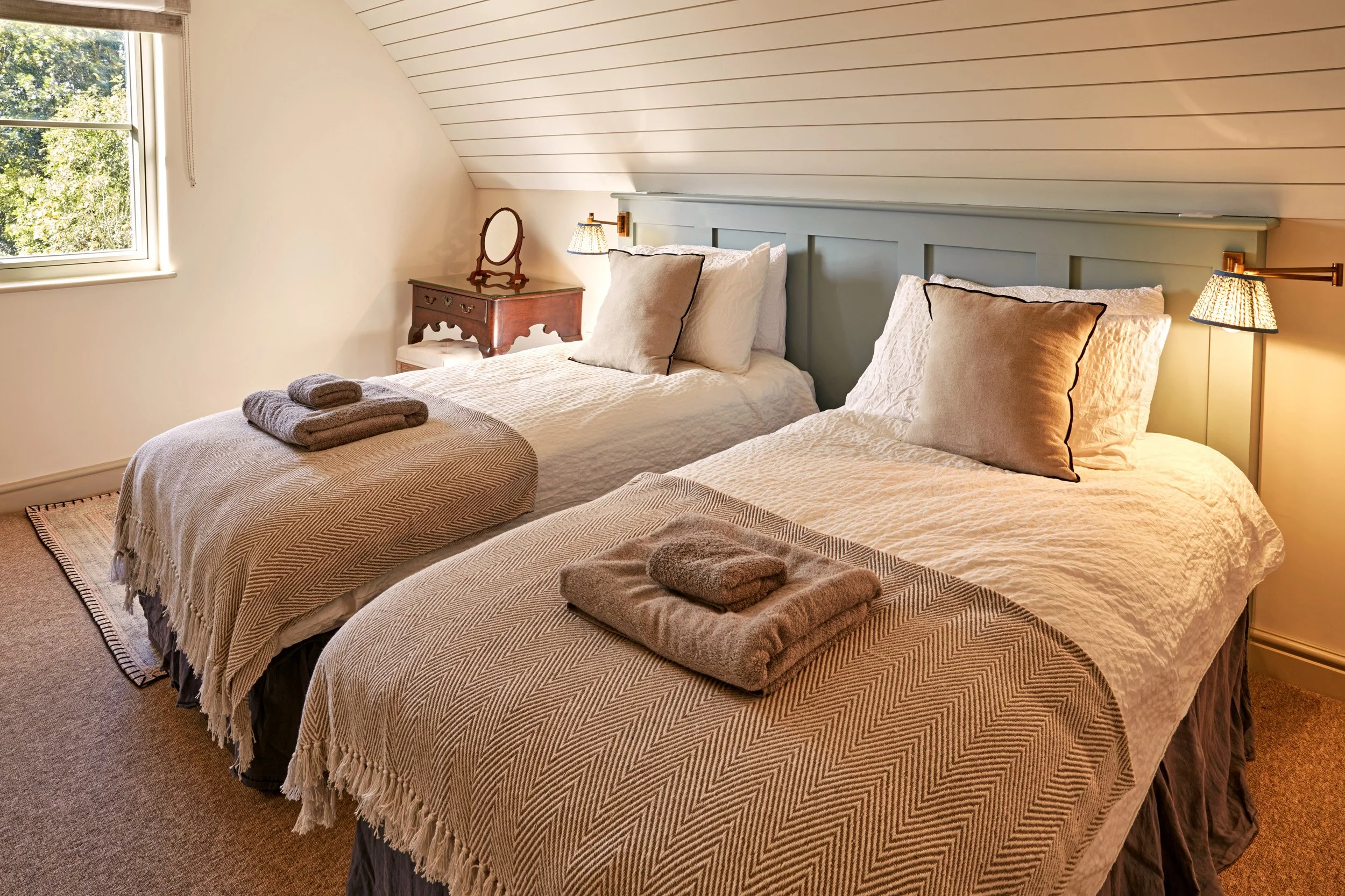A cozy bedroom with two twin beds, beige and white bedding, neatly folded towels on each bed, a small wooden bedside table with an antique mirror, and wall-mounted lamps with warm lighting. A window shows green trees outside.