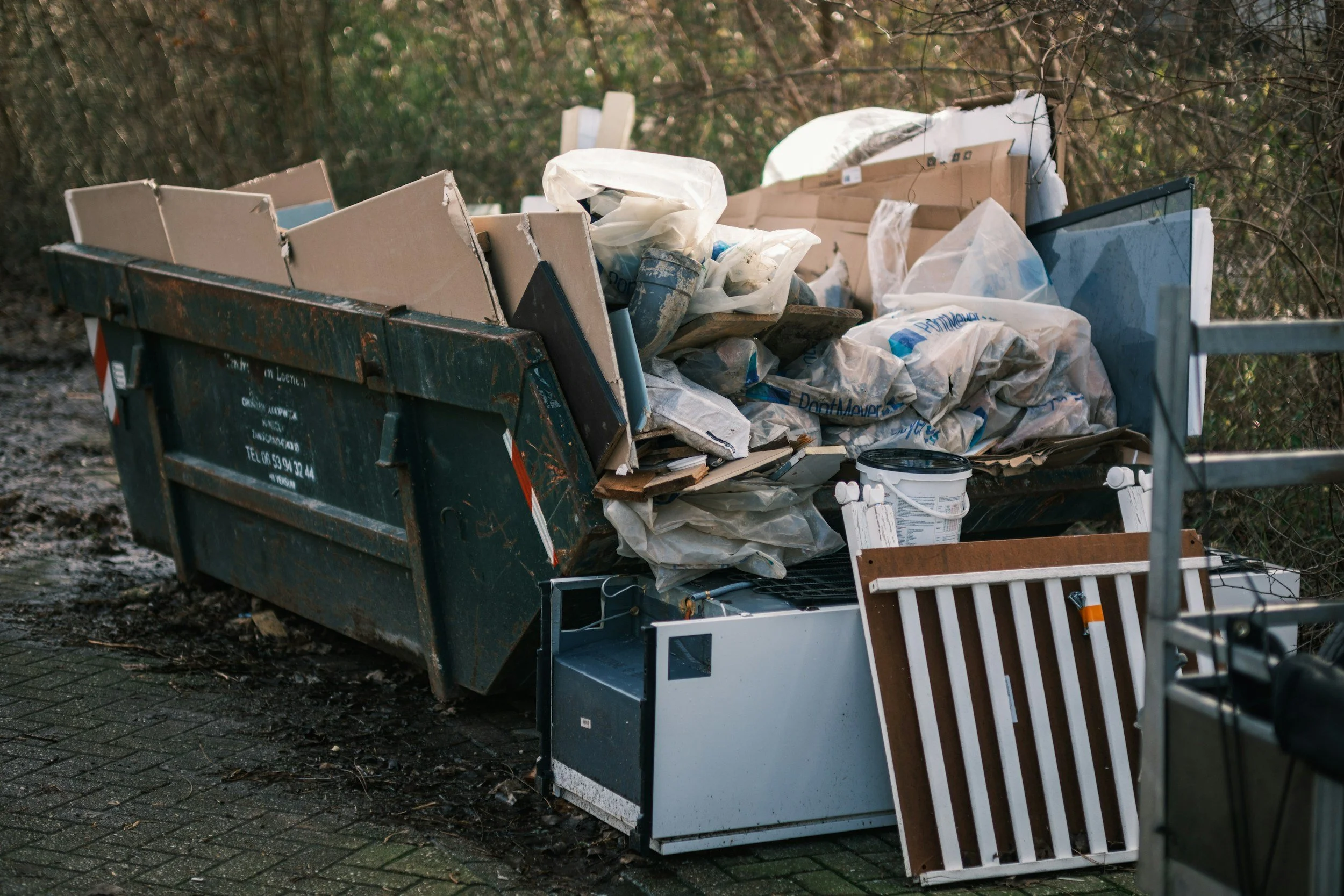 A dumpster overflowing with trash, including cardboard boxes, plastic bags, and various debris, situated on a sidewalk next to a wooded area.