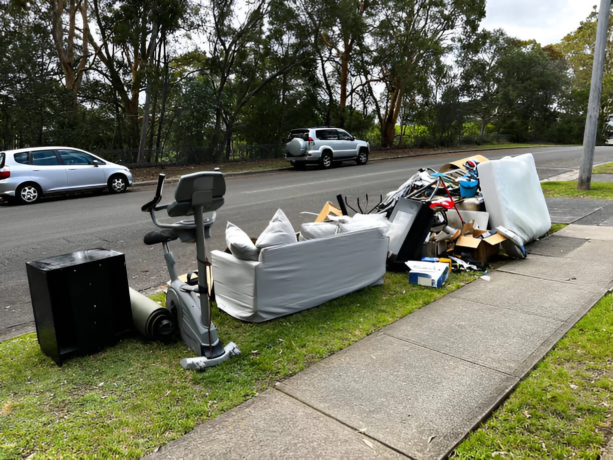 Piles of discarded furniture and household items on a sidewalk and street, including a mattress, a couch, a chair, a desk, and various boxes and debris.