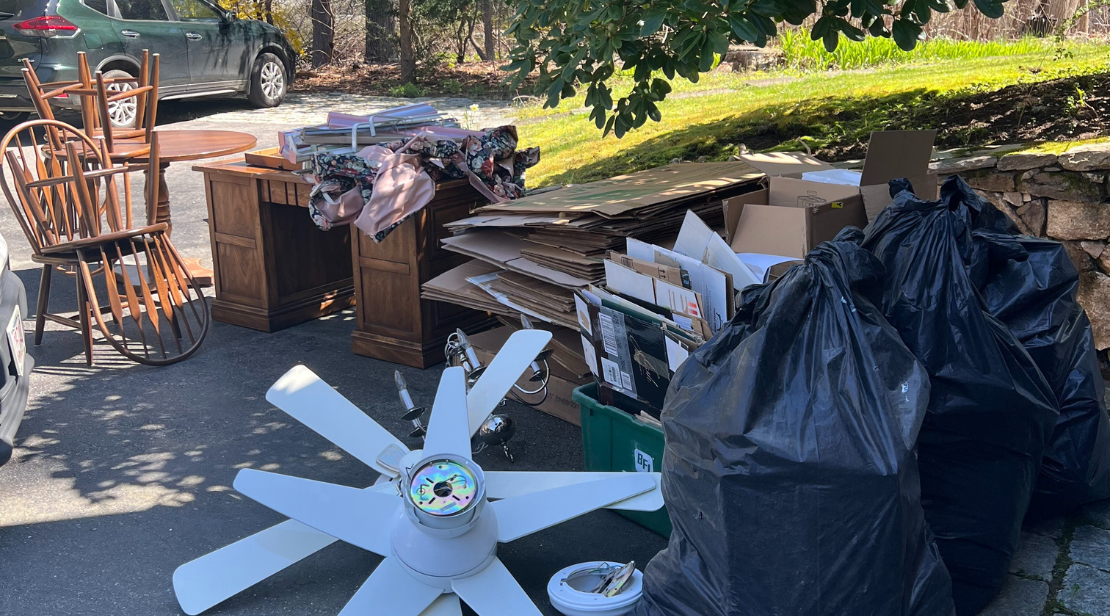 Outside scene of a garage sale with furniture, cardboard boxes, garbage bags filled with items, a ceiling fan, and a green recycling bin
