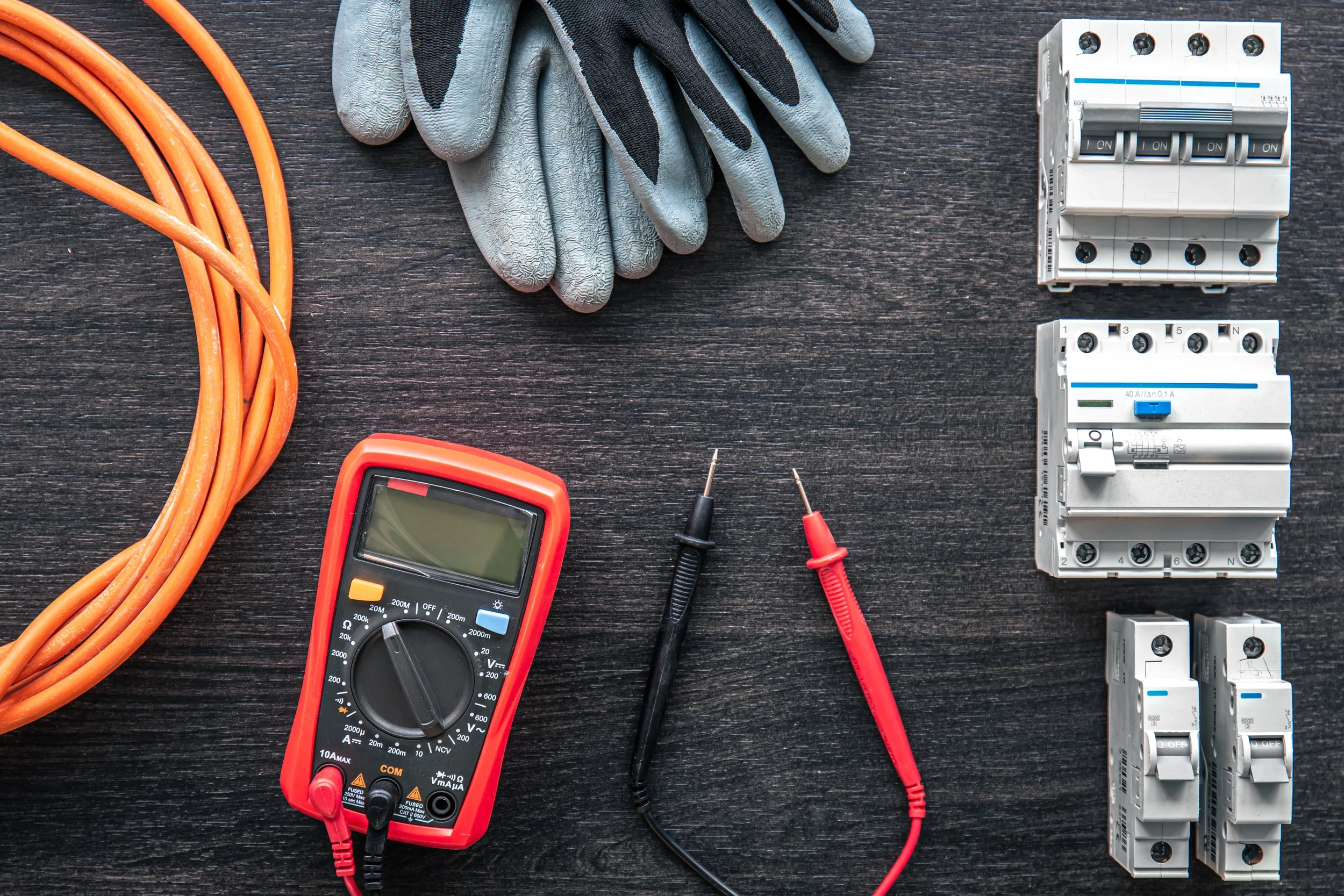 Outils pour travaux électriques, incluant un multimètre, gants de protection, disjoncteurs, câbles oranges et fils de test, disposés sur une surface en bois