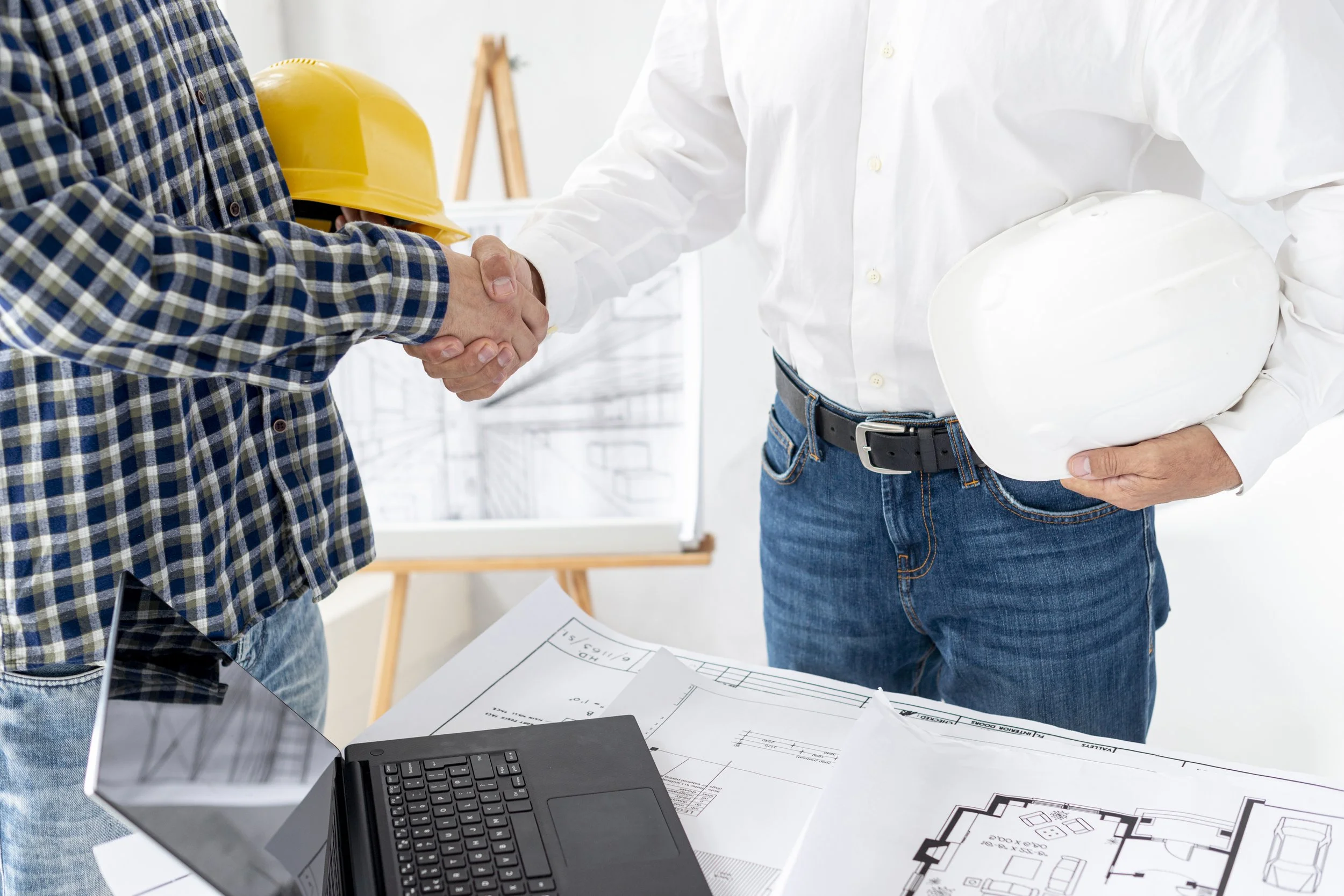 Deux personnes se serrent la main, une d'elles porte un casque de chantier jaune et un vêtement à carreaux, tandis que l'autre porte une chemise blanche et tient un casque blanc. Il y a des plans et un ordinateur portable sur une table, dans un environnement d'atelier ou de bureau de construction.