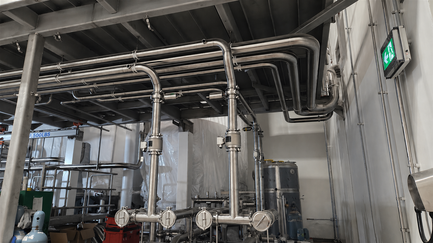 Les tuyaux en acier inoxydable forment un réseau complexe dans une installation industrielle, avec un panneau de sortie d'évacuation d'urgence vert avec des flèches indiquant la direction.