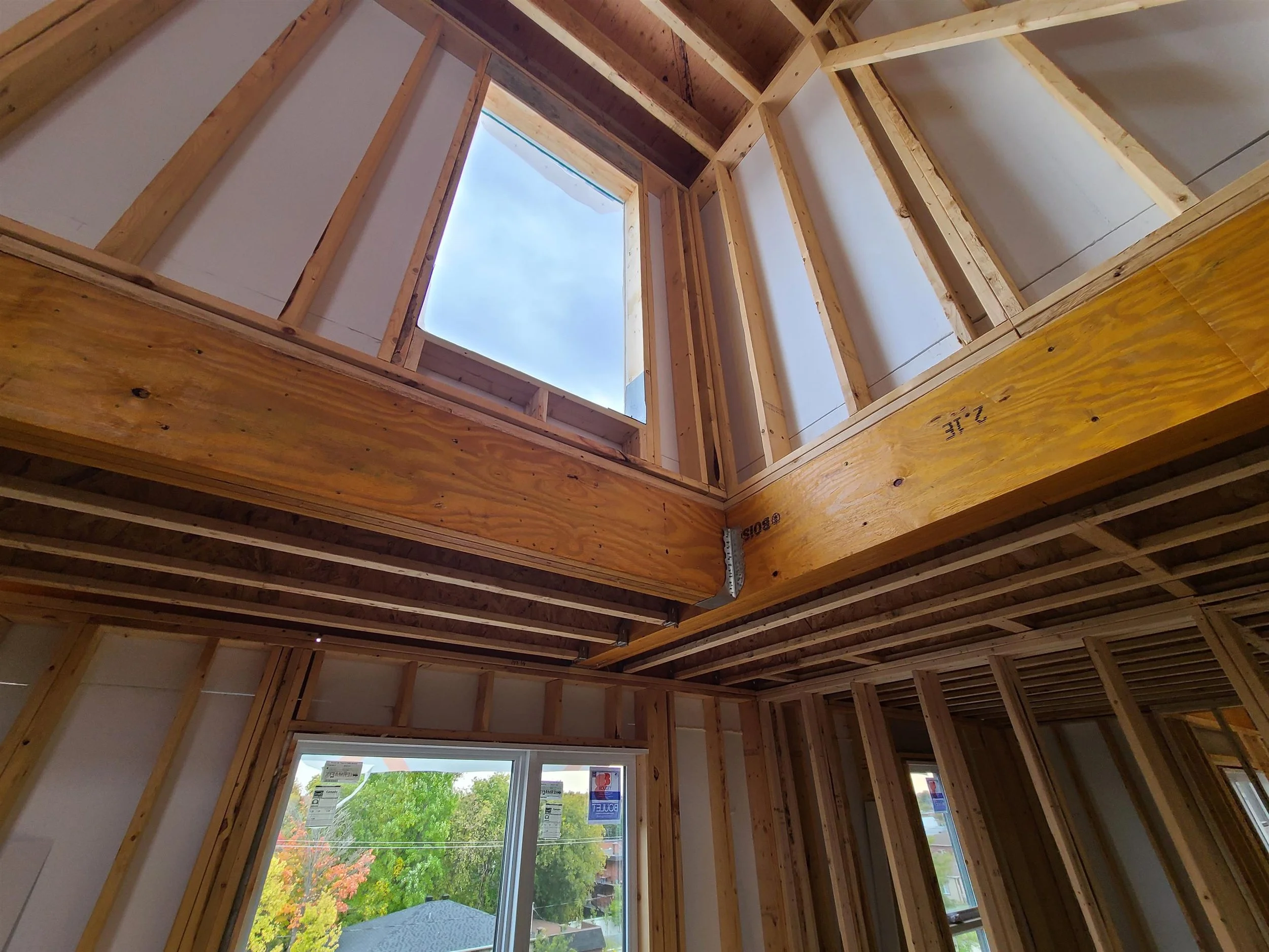 Construction intérieure d'une maison avec des murs en bois et un plafond partiellement ouvert laissant entrer la lumière naturelle, vue vers l'extérieur avec des arbres et des maisons voisines.