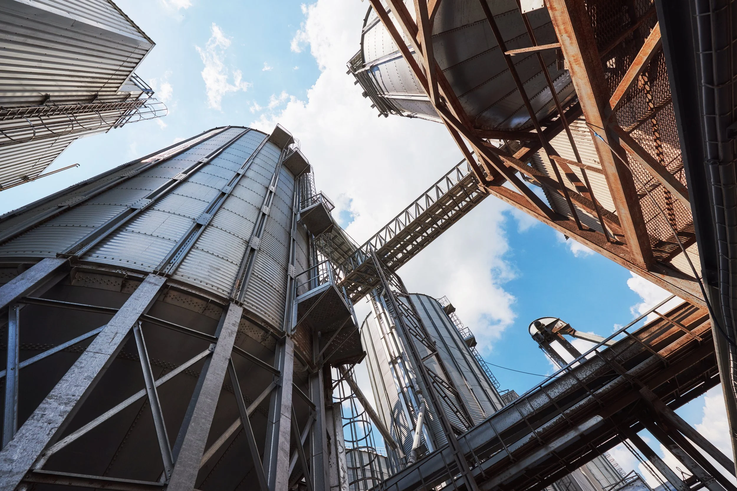 Vue d'en bas d'une usine avec des silos en métal et des structures en acier, le ciel en arrière-plan avec quelques nuages.