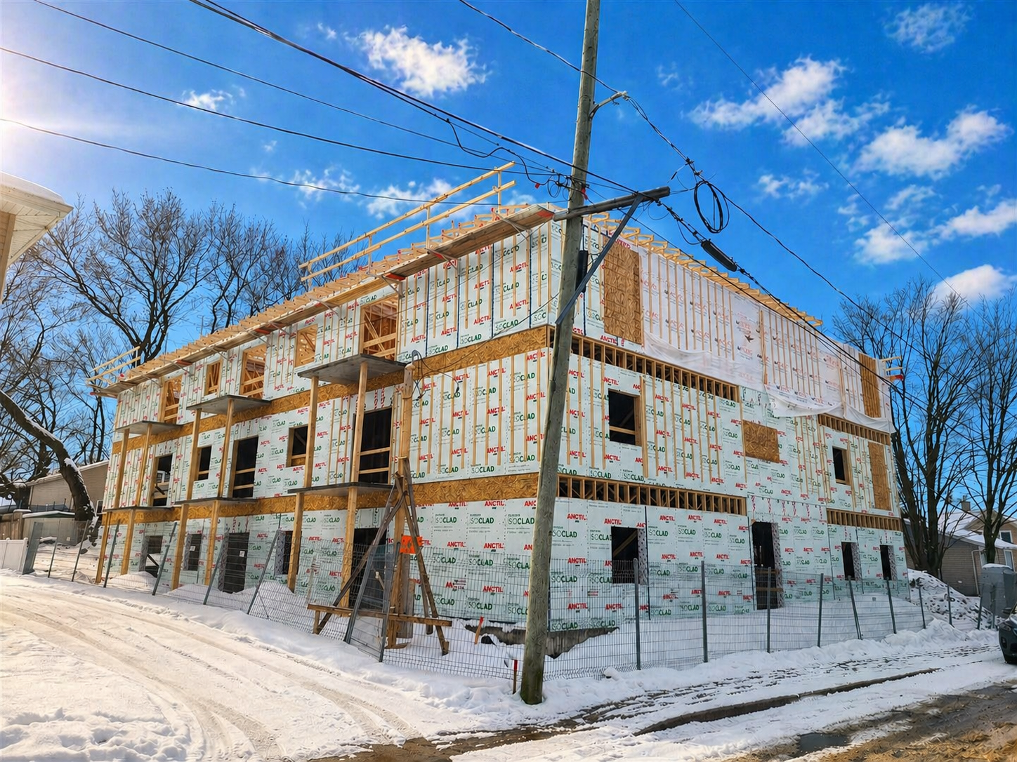 Construction d'un bâtiment résidentiel en cours, structure en bois et isolation enroulée, neige au sol, ciel bleu avec quelques nuages, arbres sans feuilles en arrière-plan.
