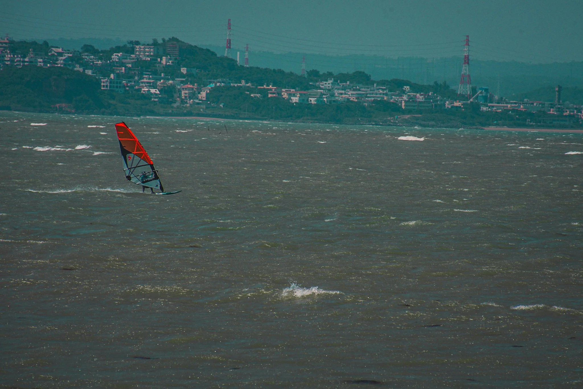 A windsurfer on choppy ocean waters with a city hillside in the background and several transmission towers.