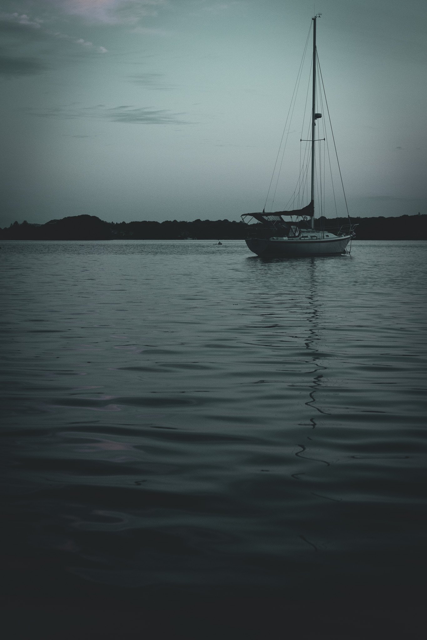 A sailboat floating on calm water near a distant shoreline under an overcast sky.