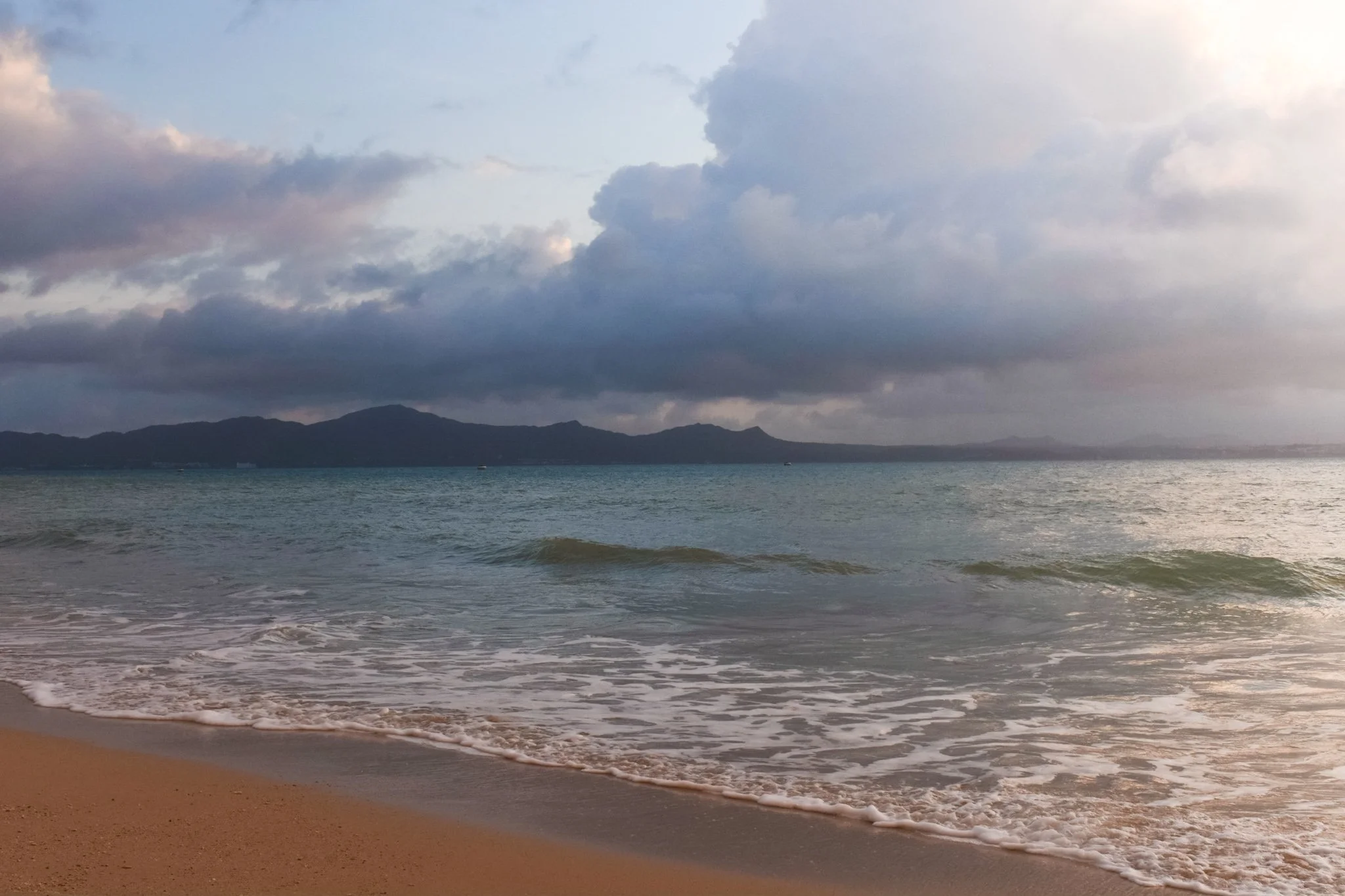 A beach scene with waves gently hitting the sandy shore, distant mountains under a cloudy sky.