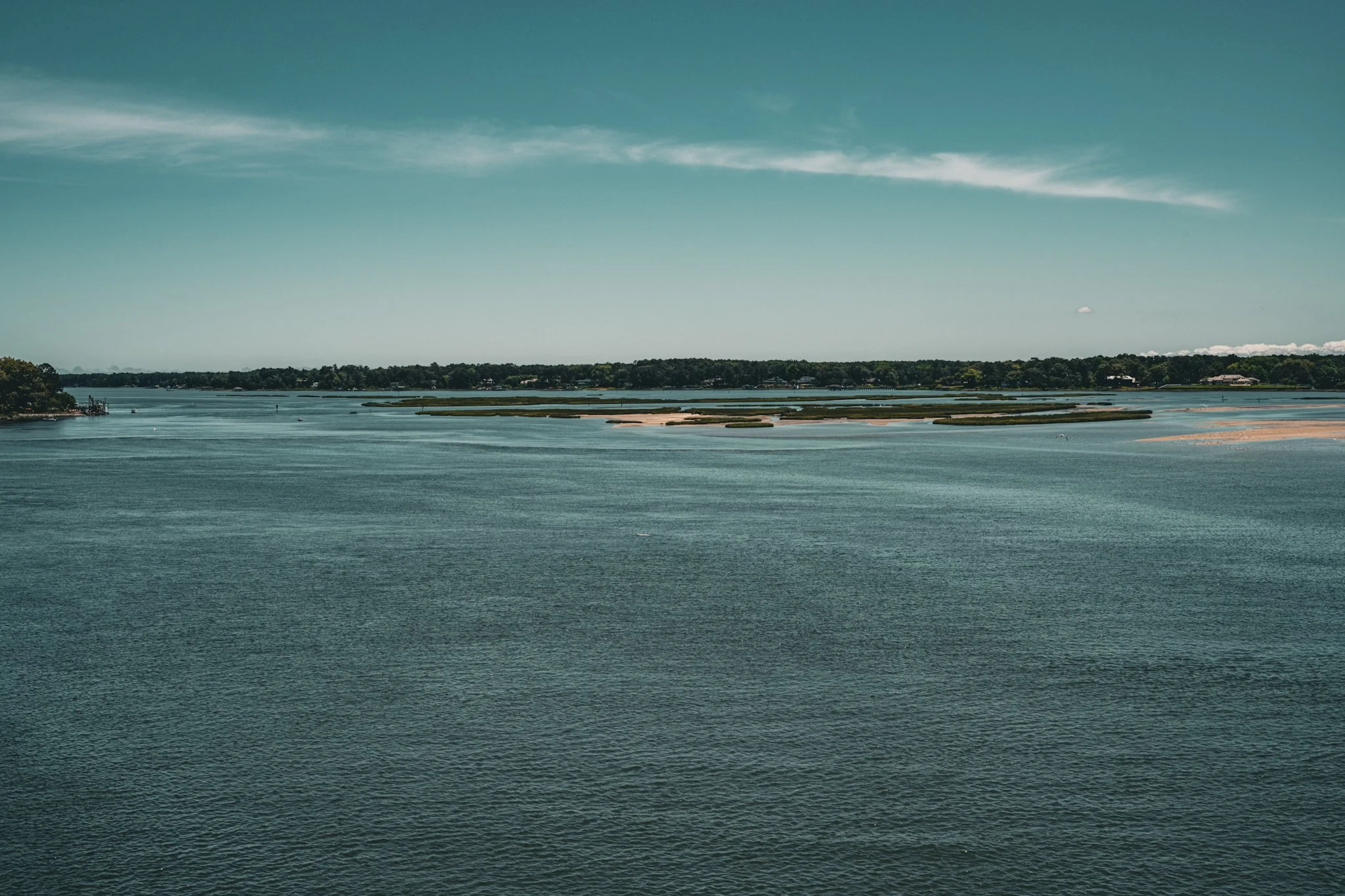 A calm body of water with small islands and a distant tree-lined shore under a partly cloudy sky.