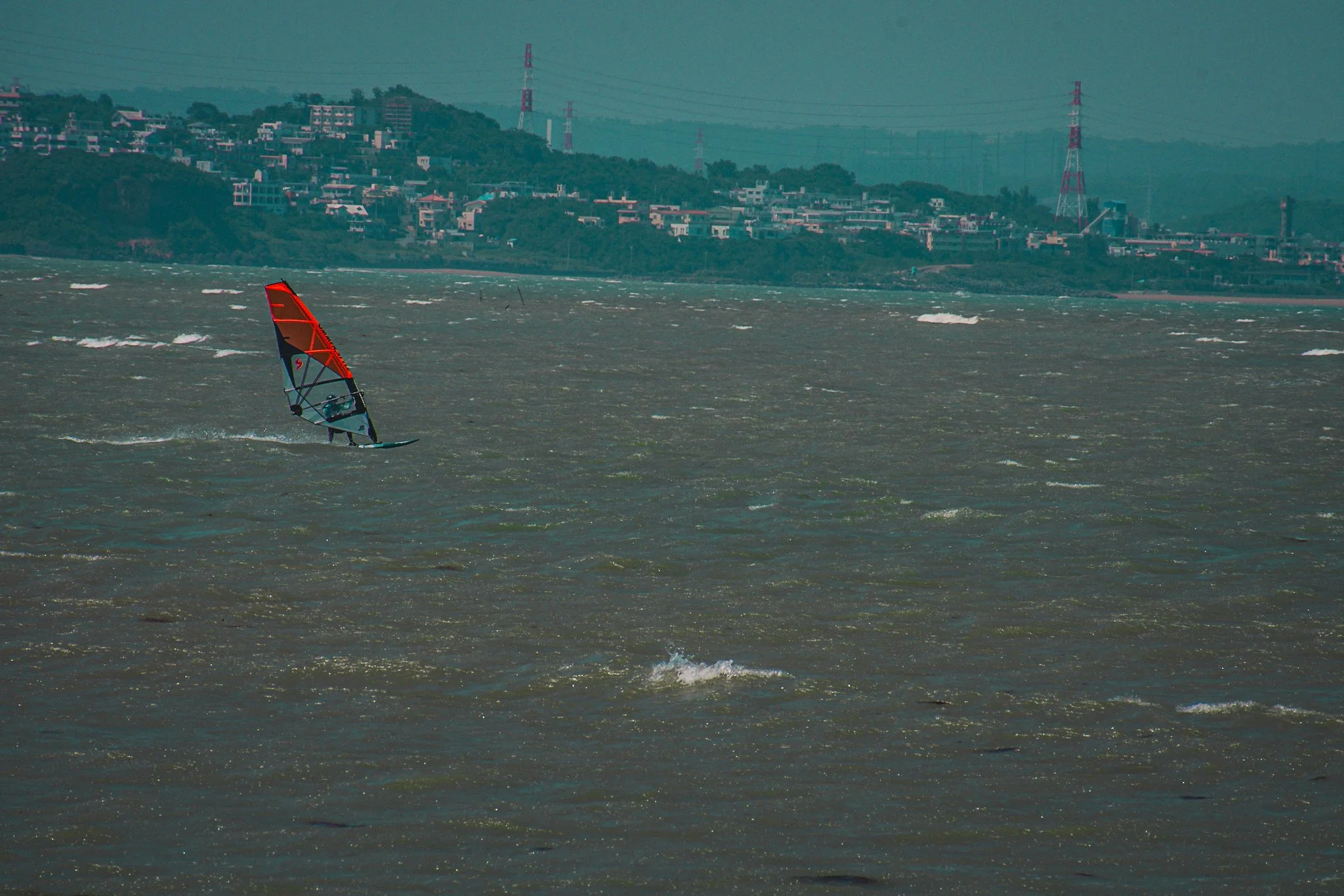 A person windsurfing on rough water near a coastal area with houses and transmission towers in the background.