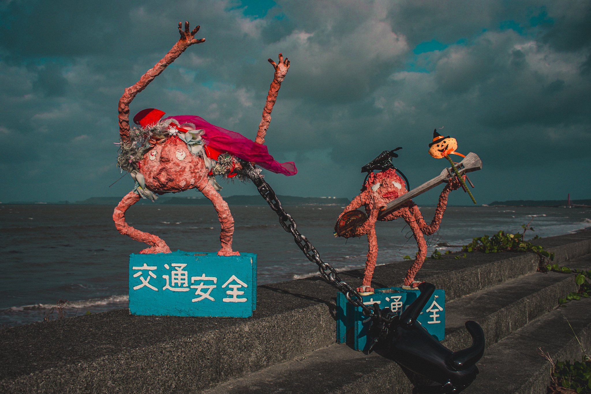 Decorative sculpture of a crab with long, bent legs and claws, adorned with Halloween-themed accessories, positioned near a body of water with signs in Japanese and a cloudy sky in the background.