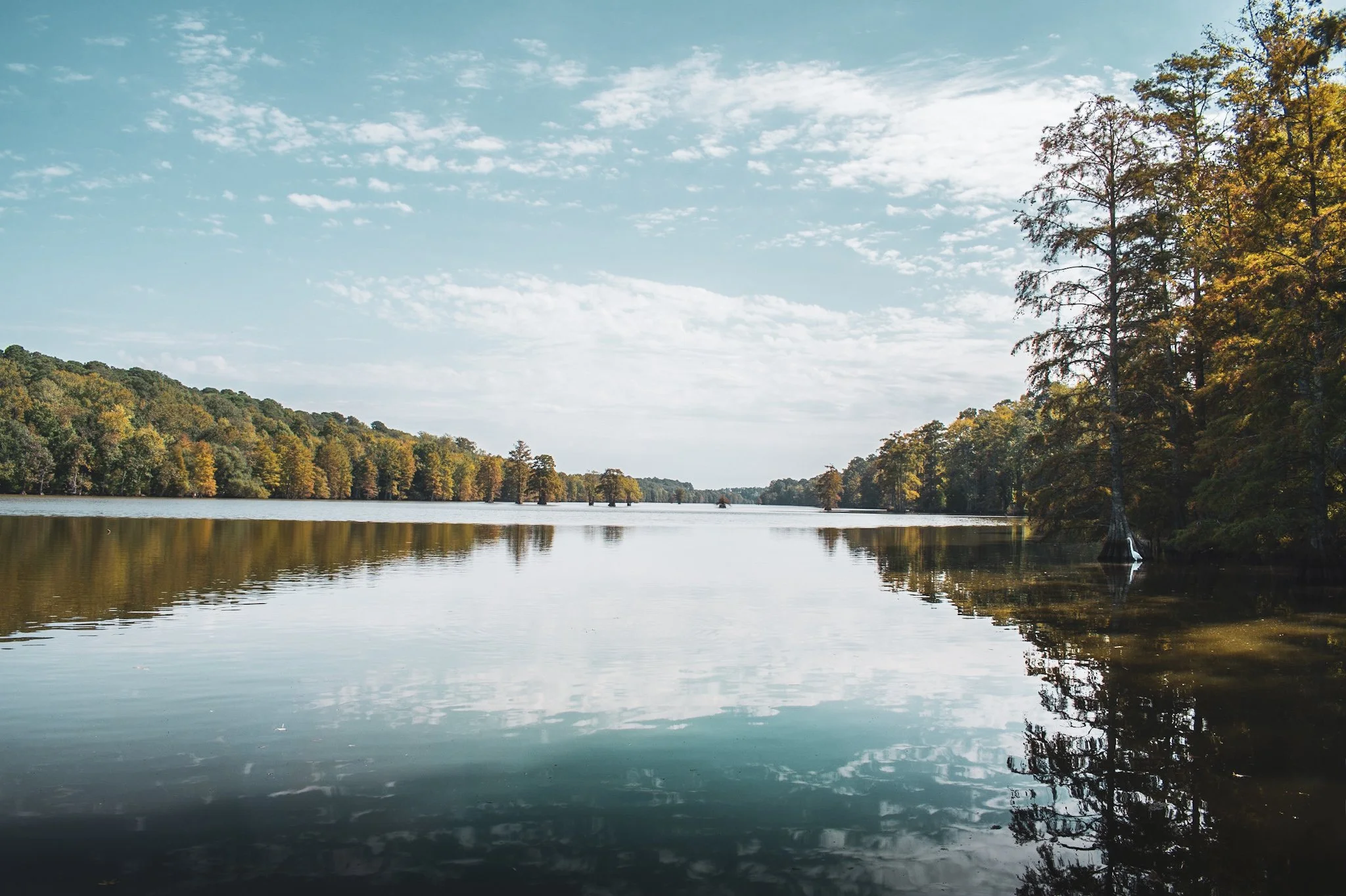 Calm river surrounded by trees with autumn foliage on a clear day with a partly cloudy sky.